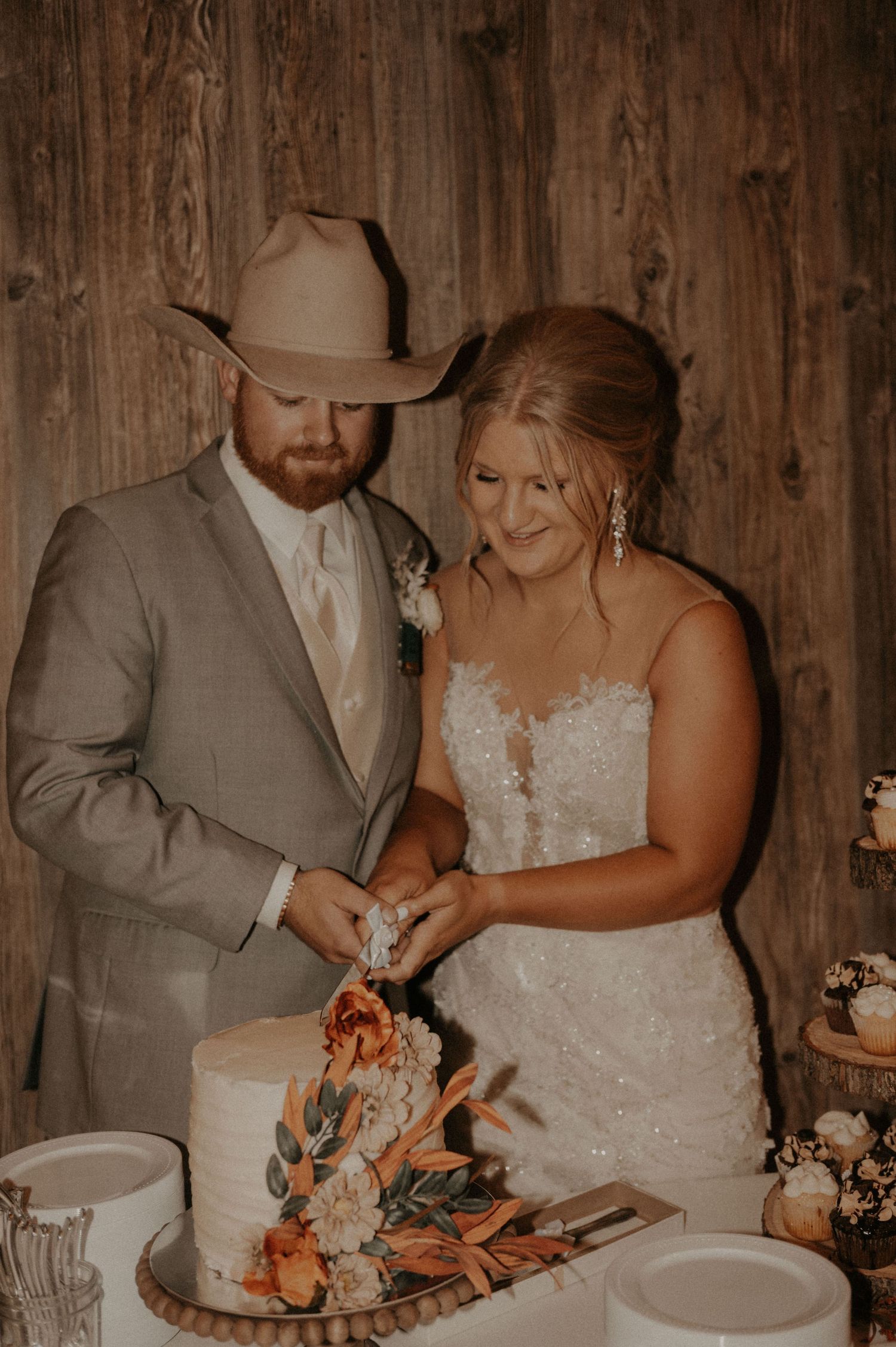 A couple in wedding attire cuts into their cake together at their rustic-themed reception.
