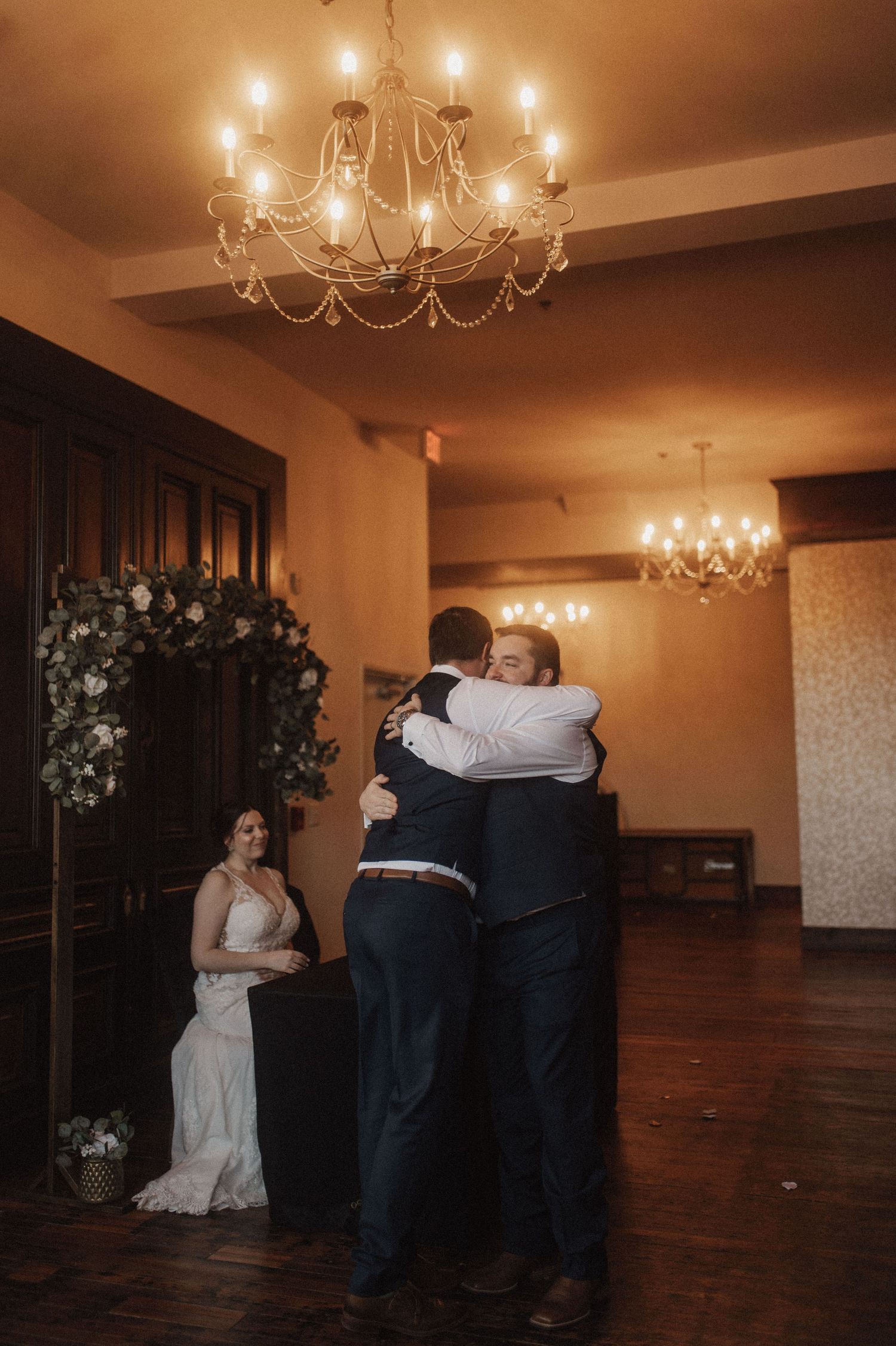 Wedding party gathers in dimly lit reception hall with elegant chandeliers.