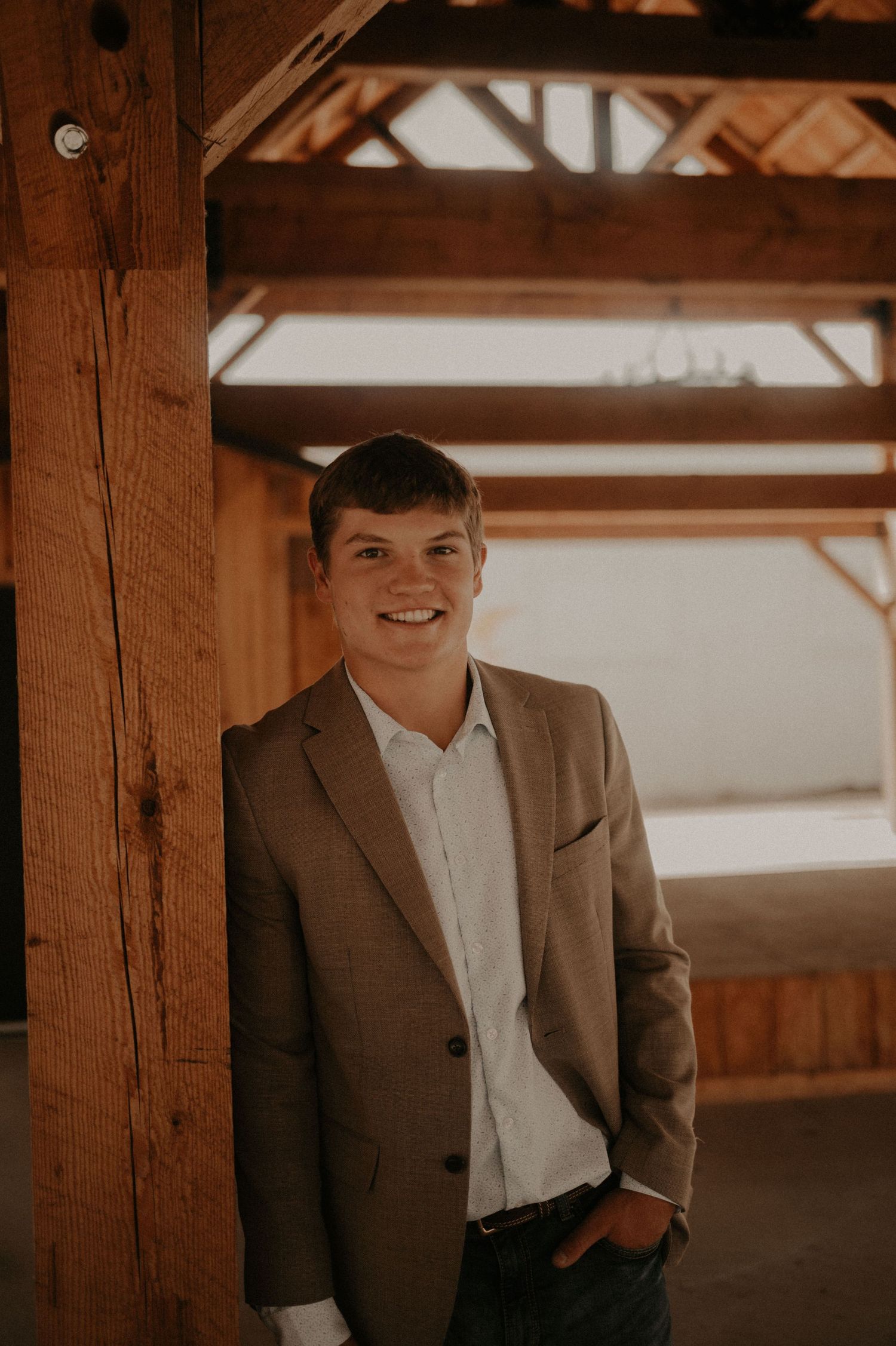 Professional portrait of someone in a brown suit jacket and white dress shirt standing against rustic wooden beams.