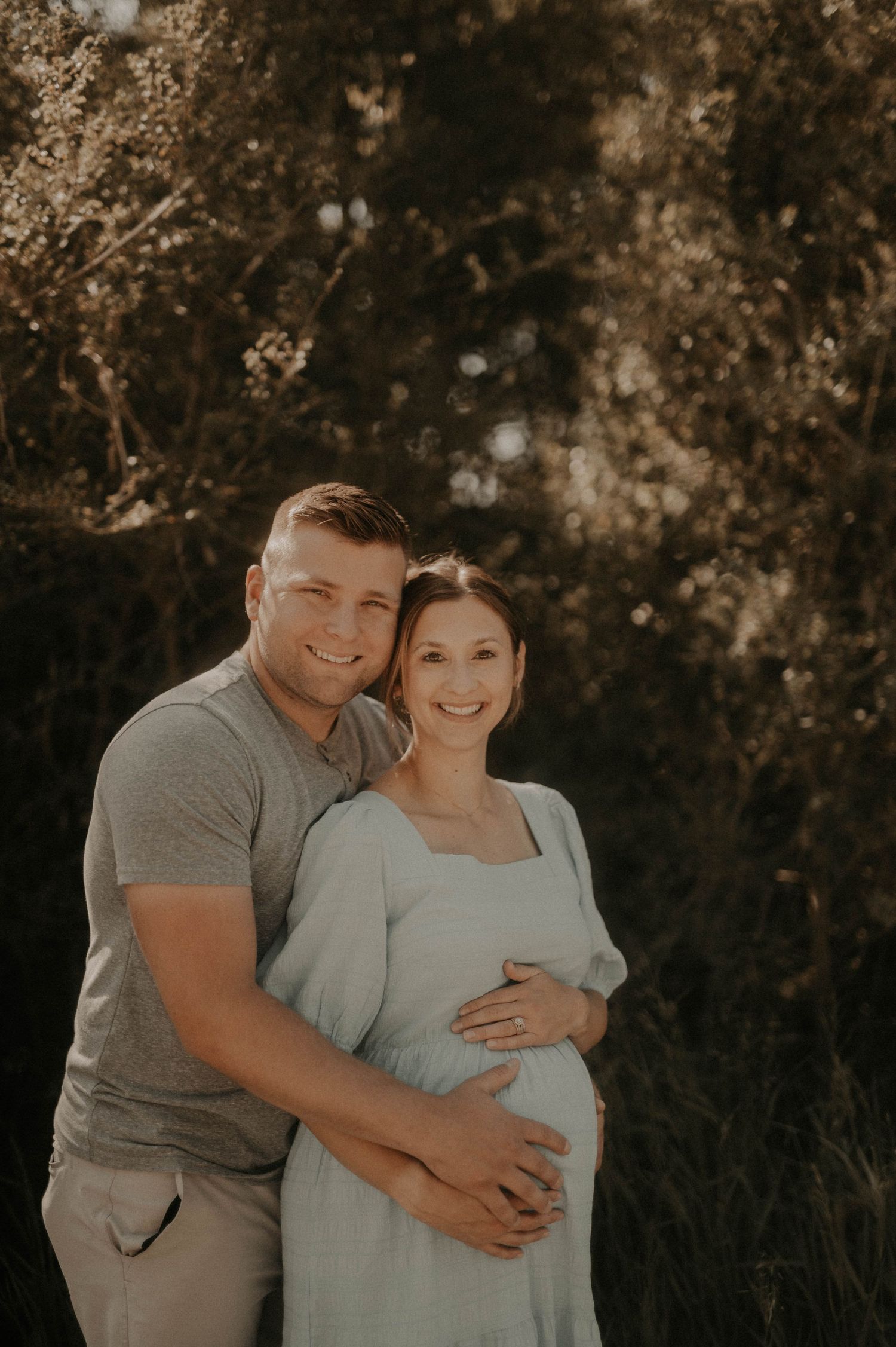A couple embraces during an outdoor maternity photoshoot in warm golden sunlight.