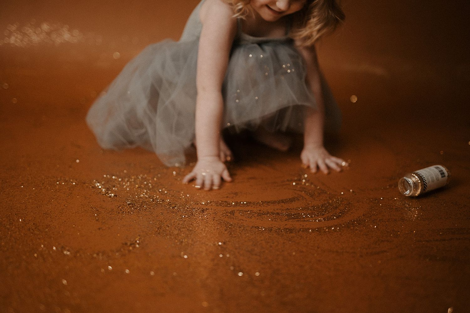 A child in a gray tutu dress sits on a rustic brown floor with glitter scattered around.