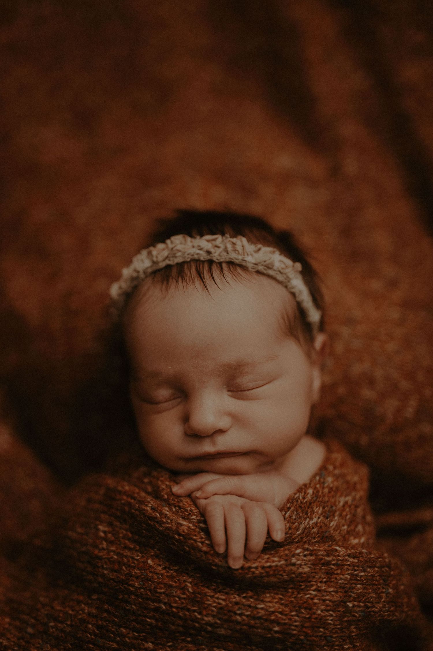 A sleeping newborn baby rests peacefully on a brown knit blanket in soft lighting.