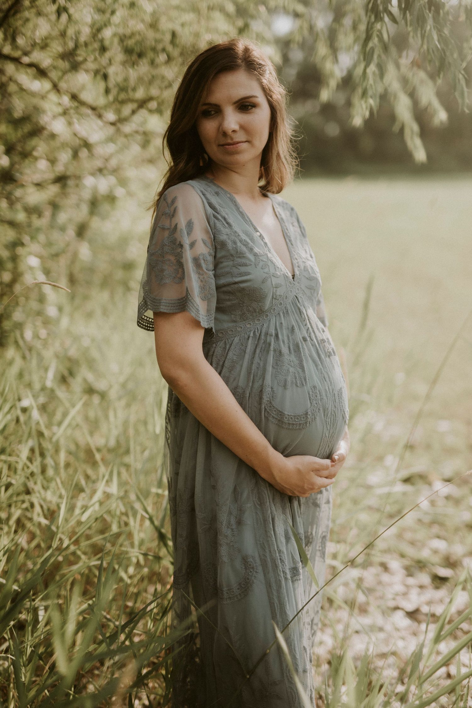 A maternity photo session in a sunlit meadow shows a pregnant woman in a flowing grey dress among wildflowers.