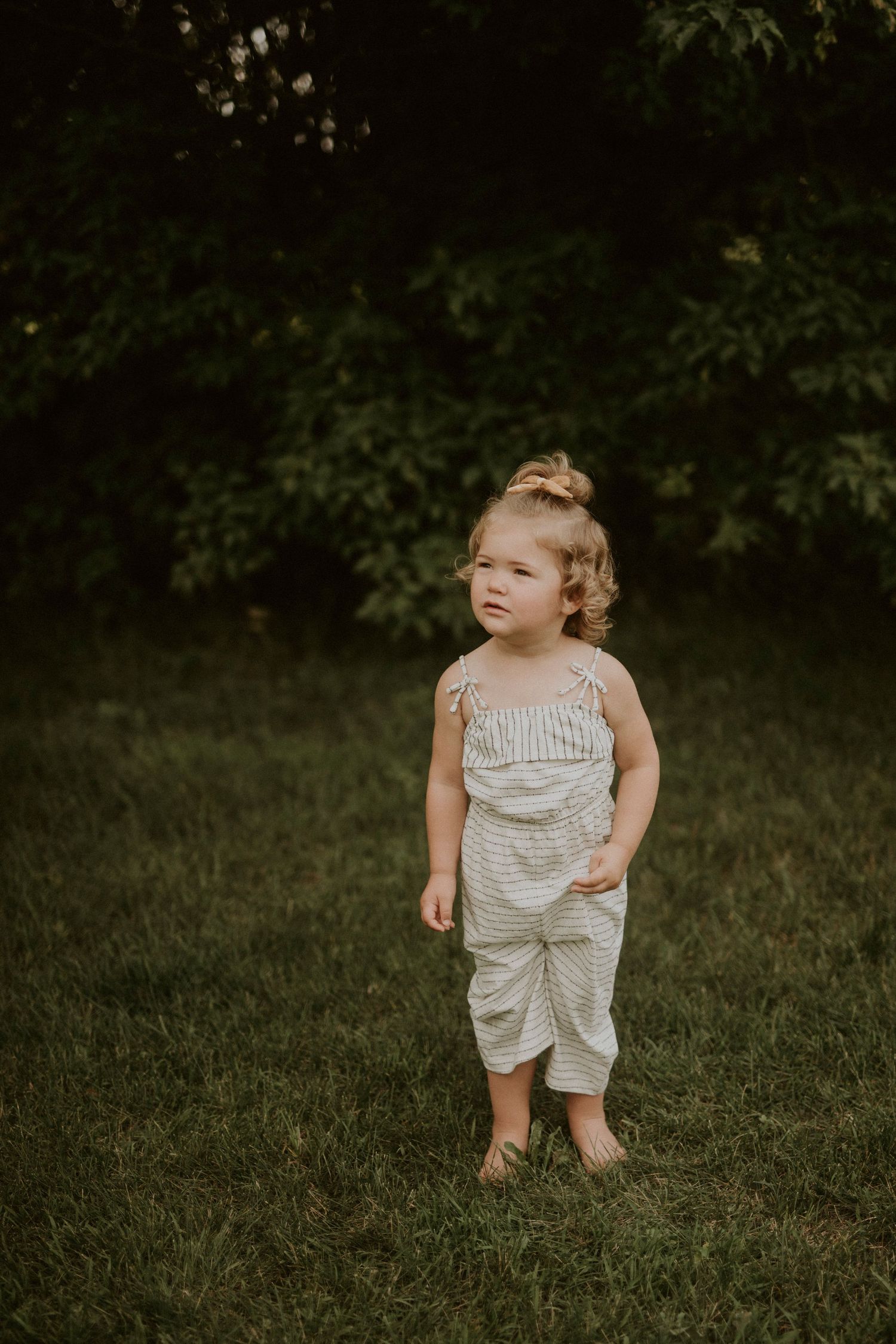 A child in striped romper explores a grassy outdoor area while looking upward on a summer day.