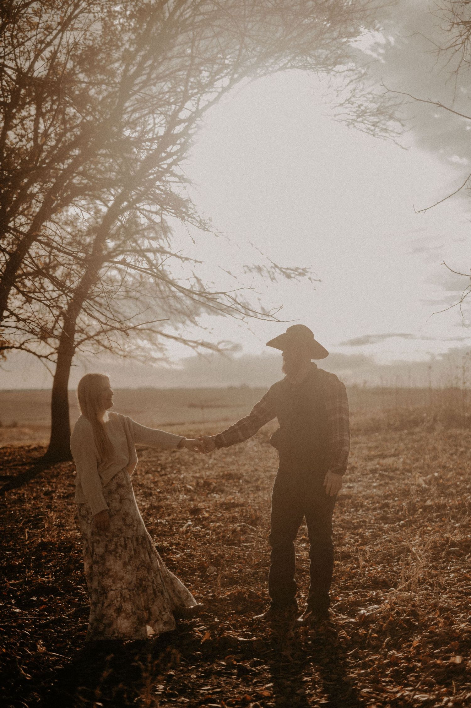 Two figures walk together through an autumn field at golden hour with trees in the background.