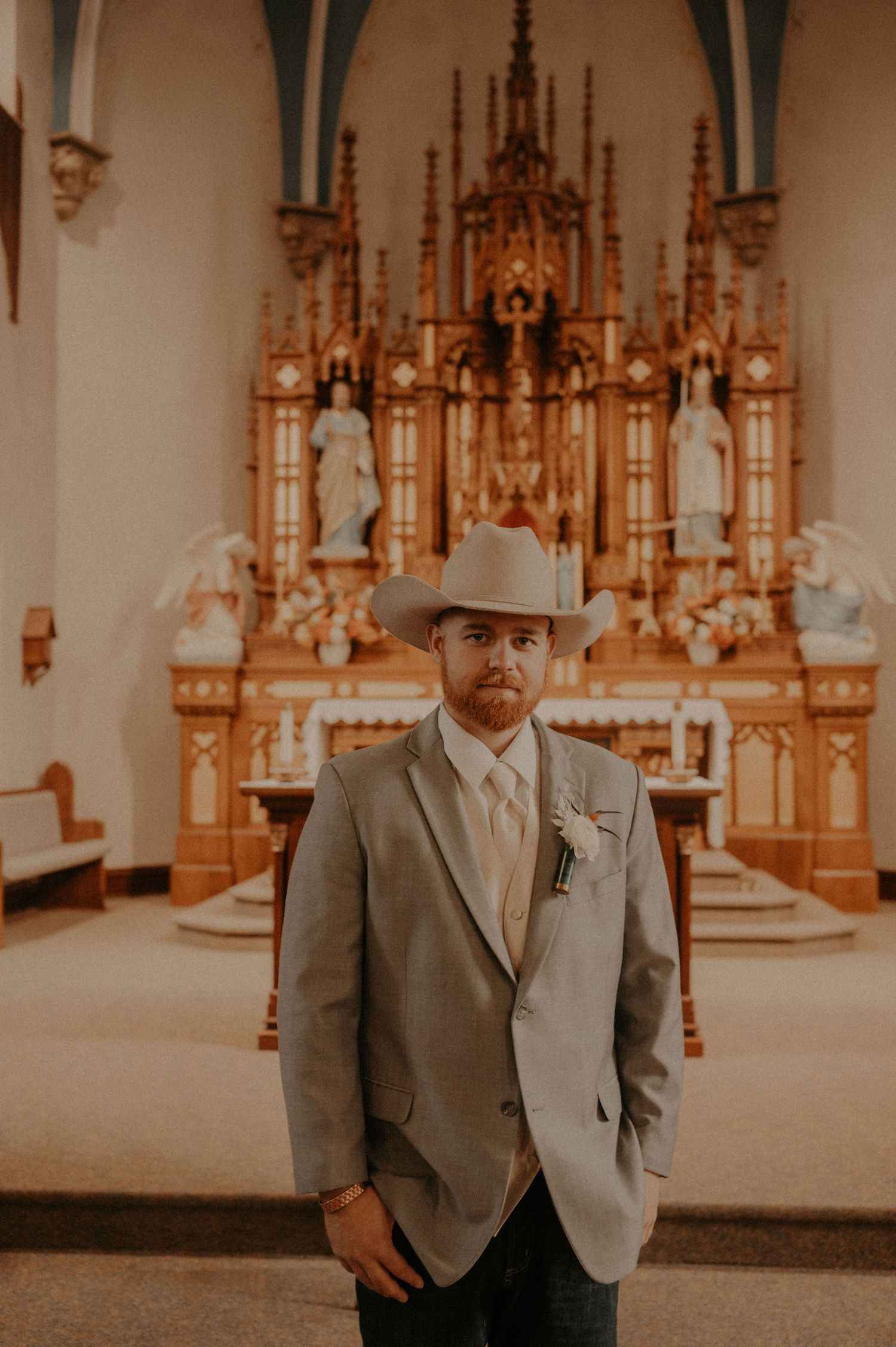 Groom in gray suit and cowboy hat stands before ornate church altar.