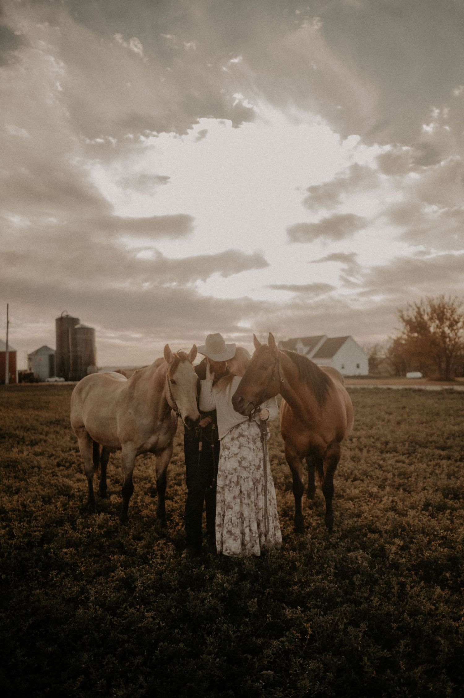 Rural engagement photo with two horses and a city skyline visible in the background.