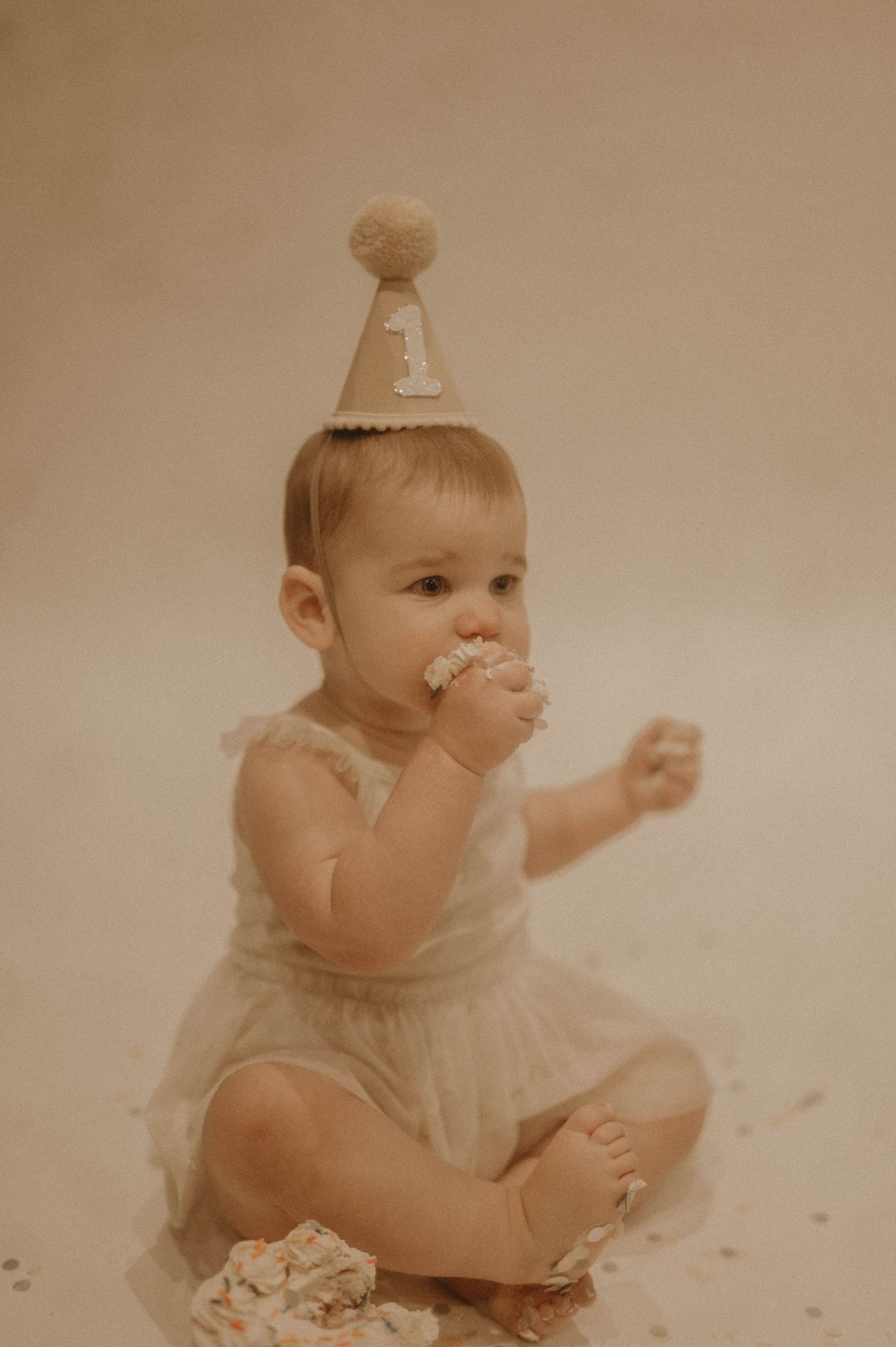 Baby wearing party hat and tulle dress sits on floor enjoying birthday cake during first birthday photo session.
