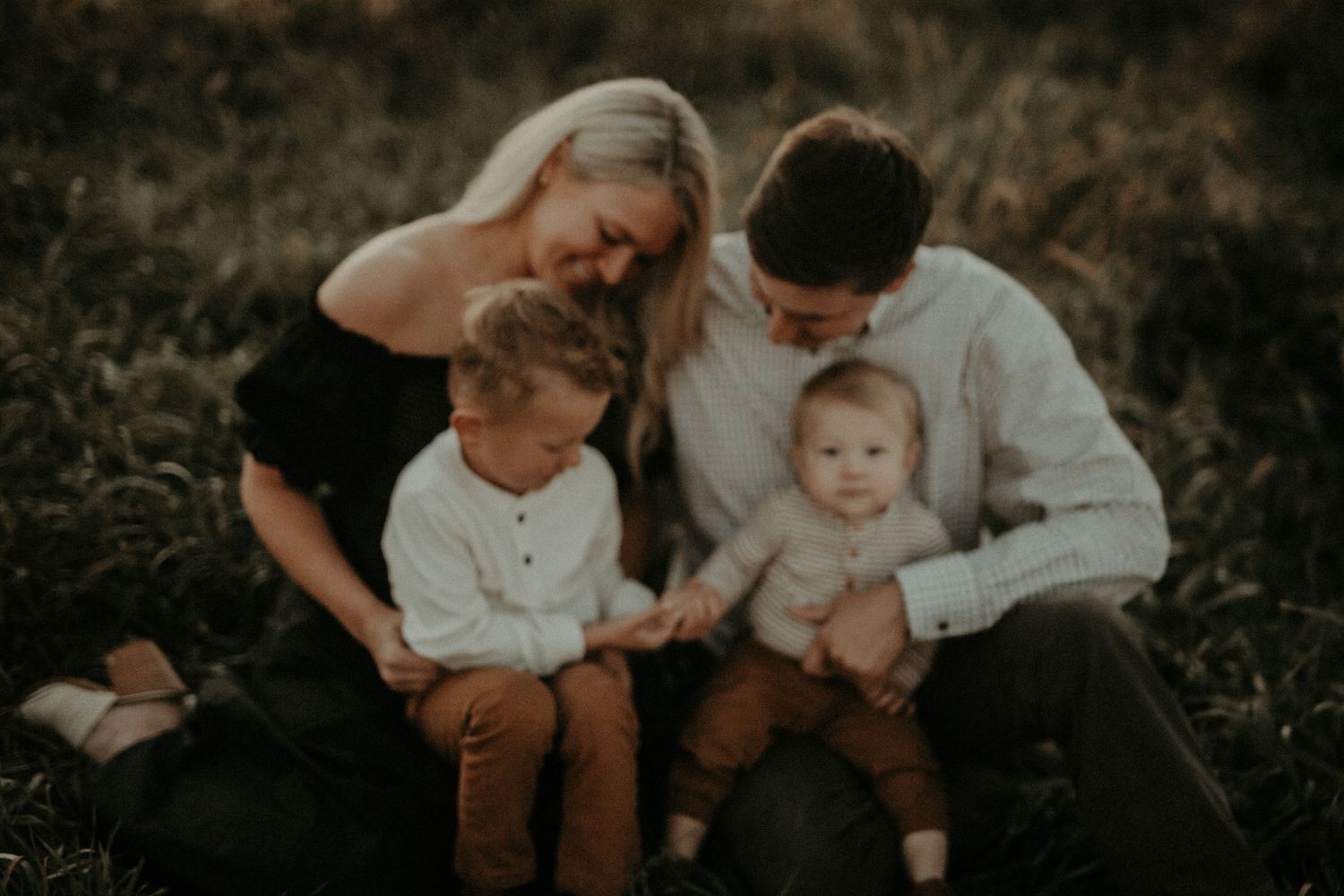A young family of four sits together in a field, with two parents and their two small children smiling warmly.