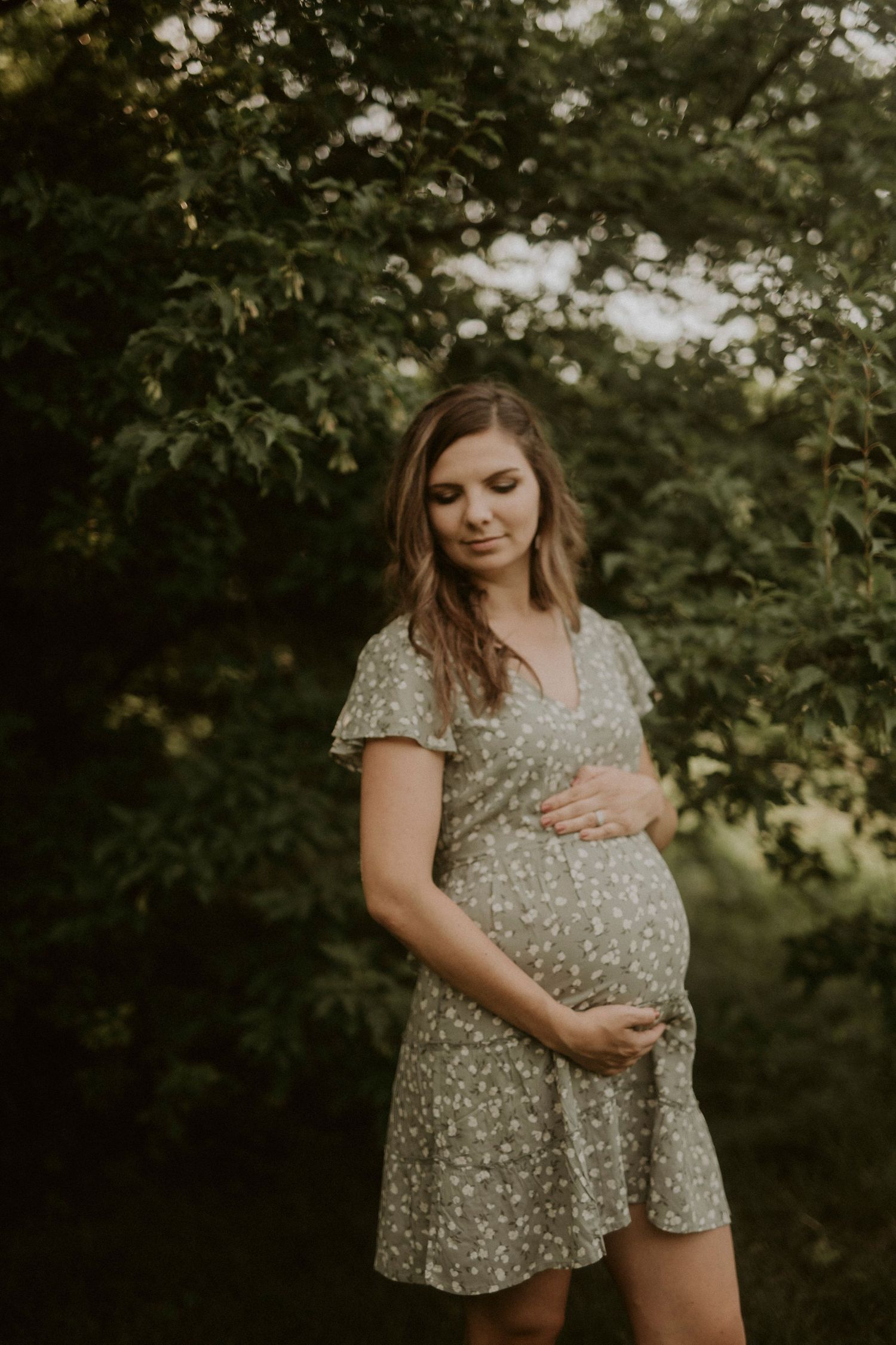 A pregnant woman in a floral dress stands in a wooded outdoor setting during golden hour.