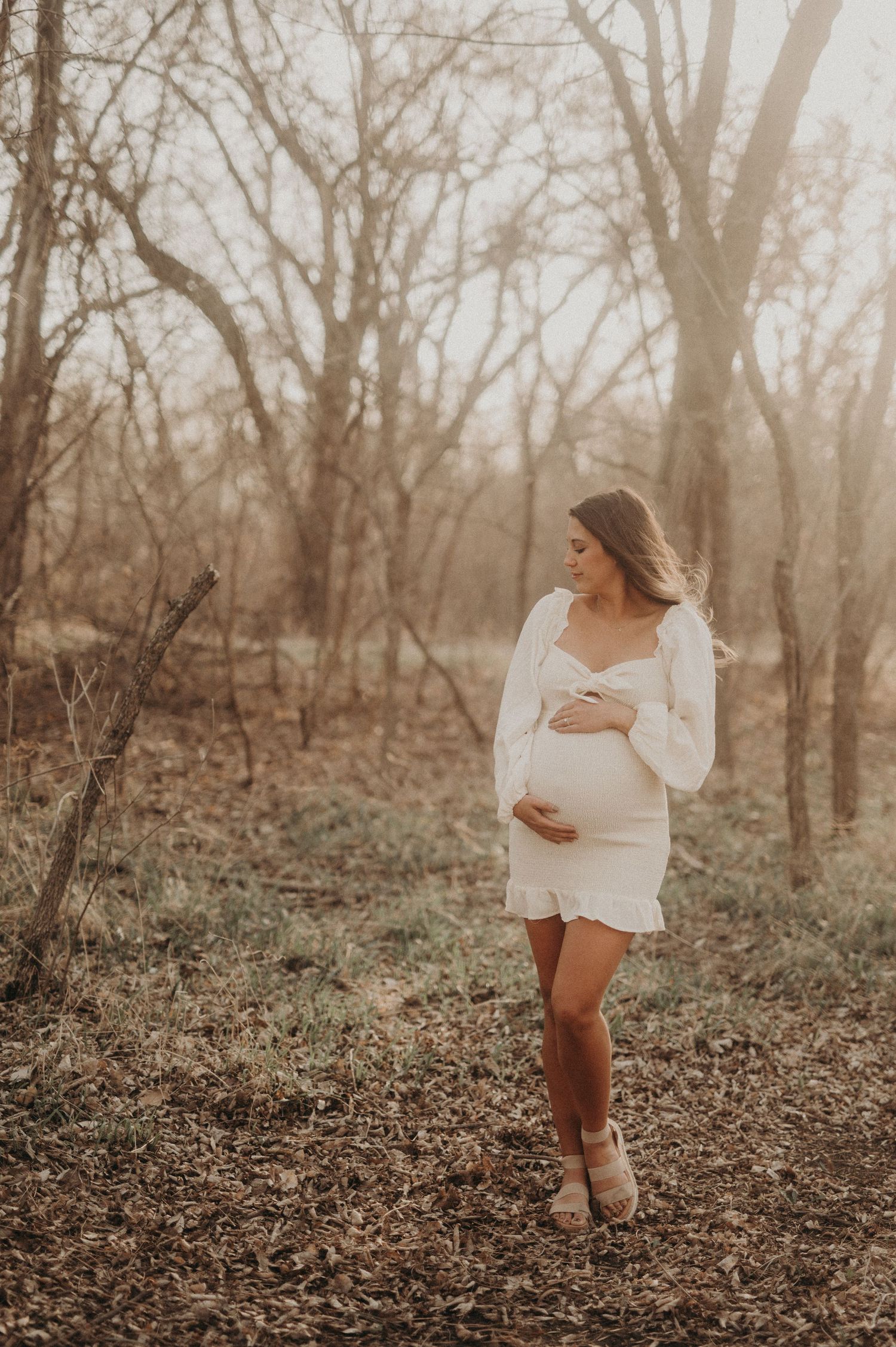 Ethereal maternity portraits of a woman in white against a wooded backdrop at sunset.