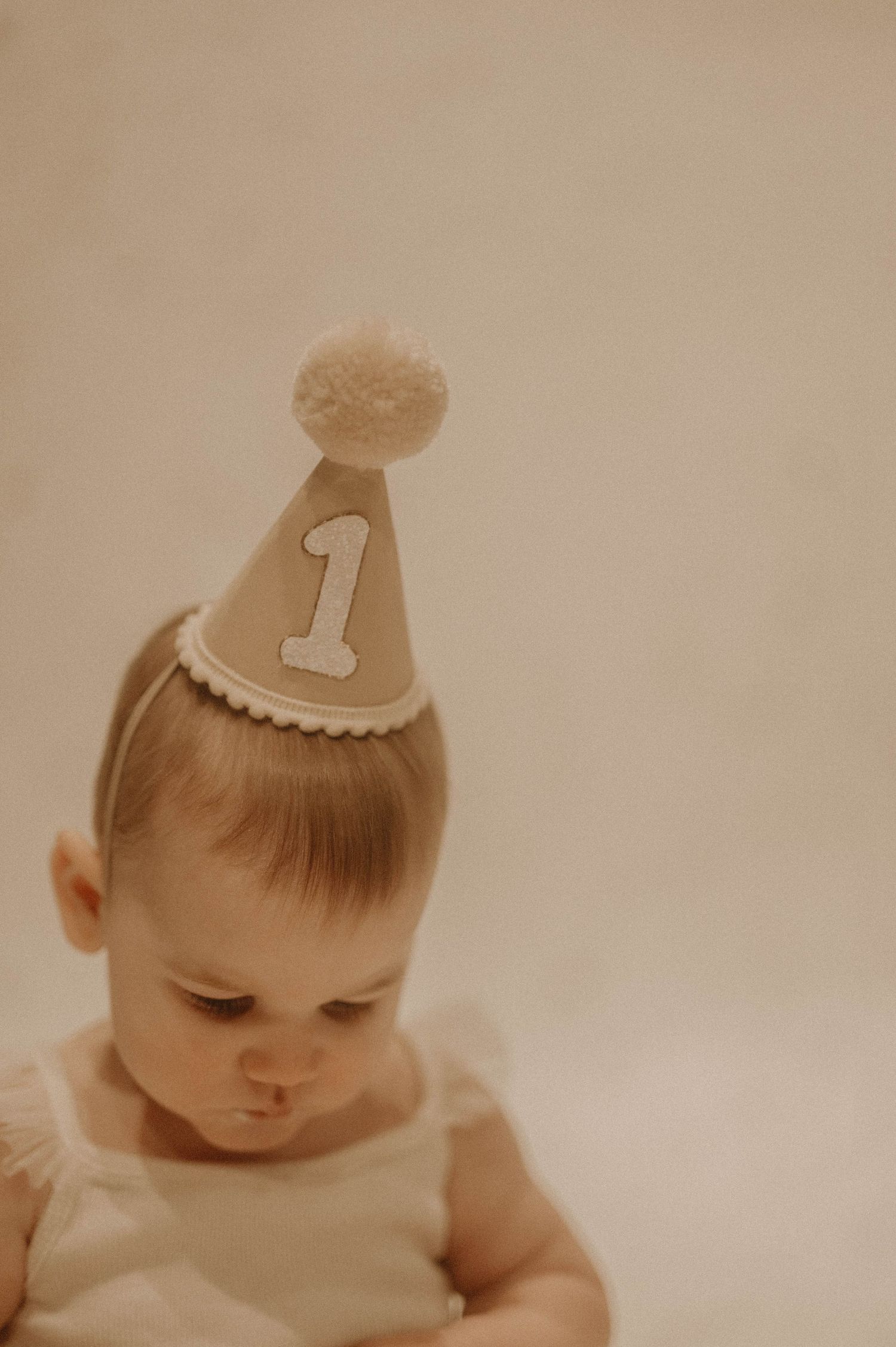 A small party hat with the number 1 sits atop a baby's head in a soft sepia-toned photograph.