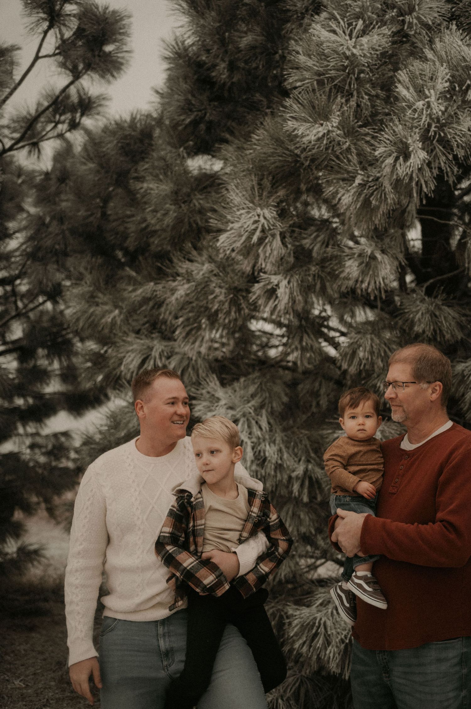 A family of three poses together in front of pine trees in a candid outdoor photo series.