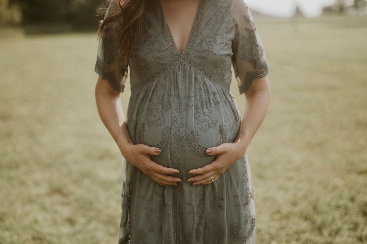 Pregnant woman in gray dress stands in grassy field during golden hour maternity photoshoot.