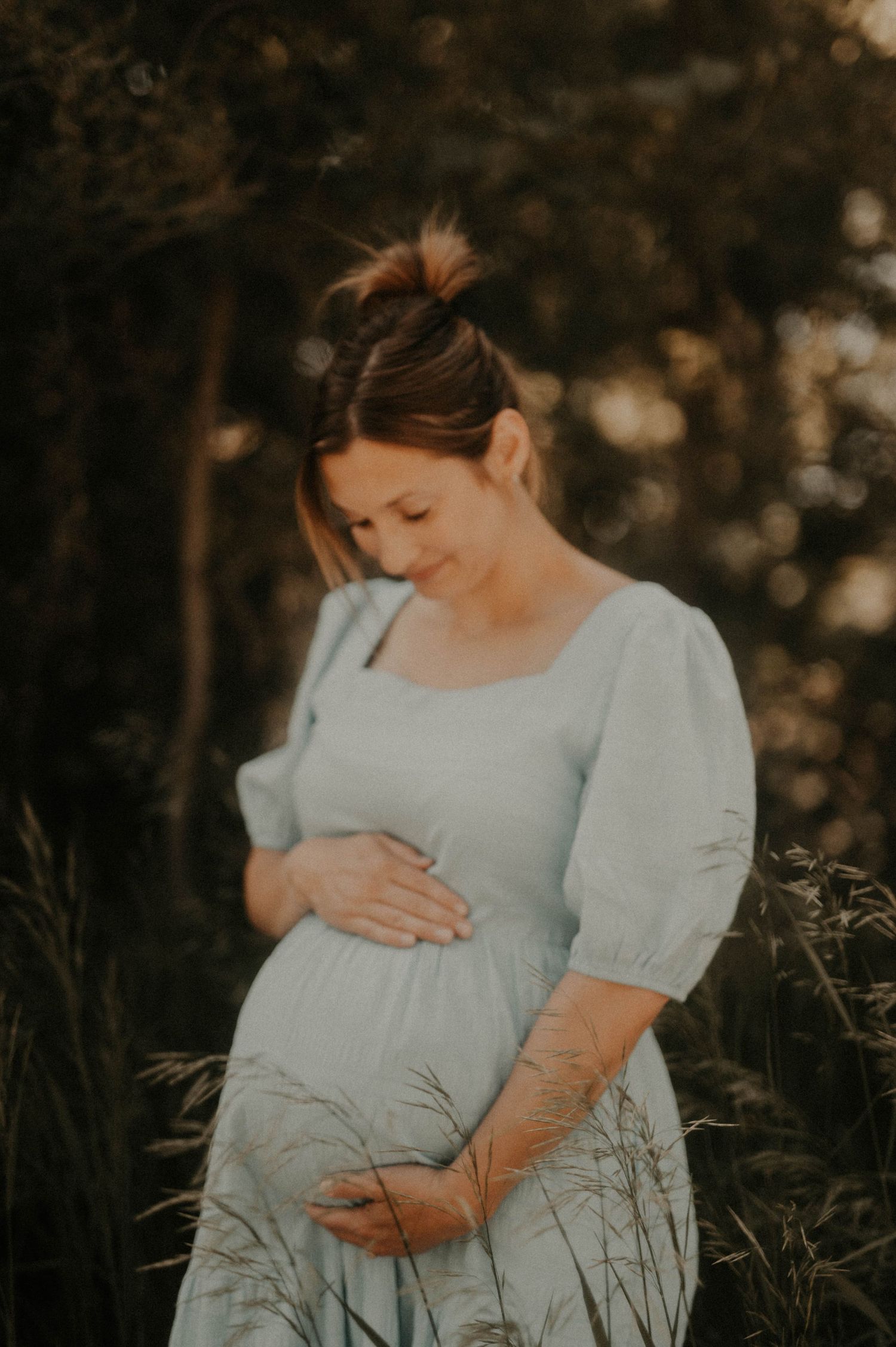 Expectant mother in a light blue dress tenderly cradles her baby bump in a dreamy outdoor maternity photoshoot at sunset.