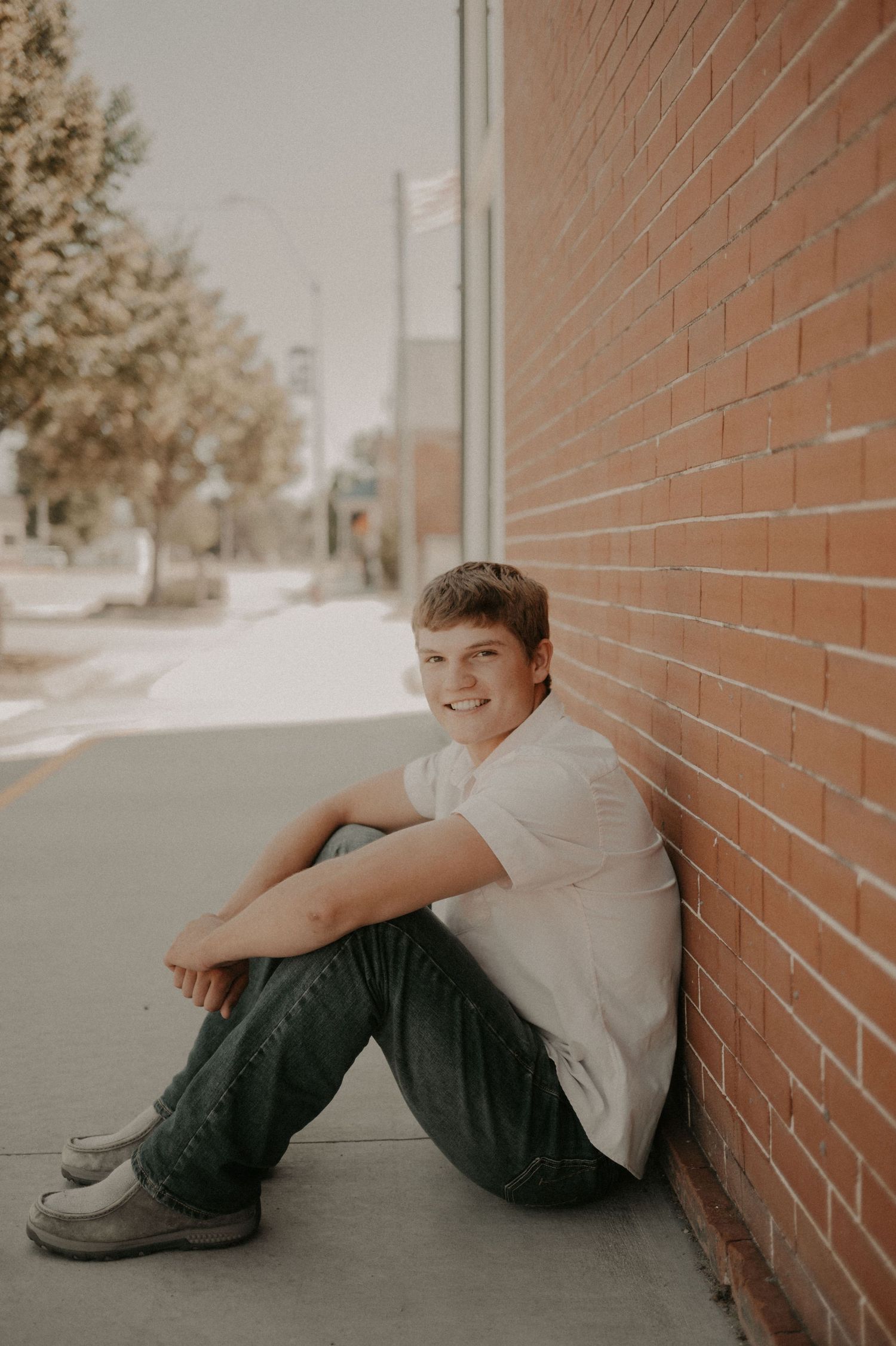 Portrait of a young person sitting against a brick wall on a sidewalk wearing a white shirt and dark pants.