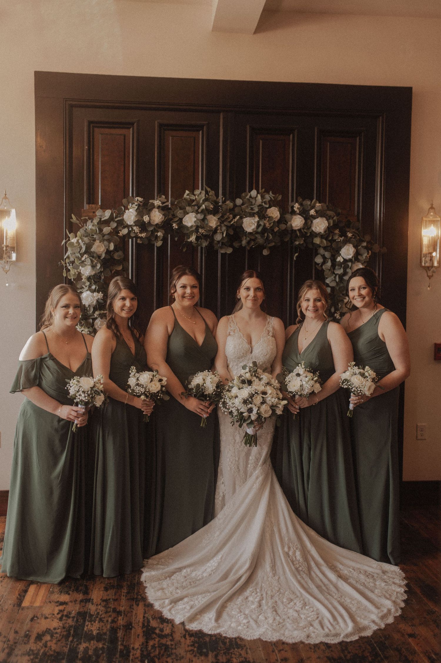 Bridesmaids in sage green dresses pose with bride under floral arch against dark wooden doors.