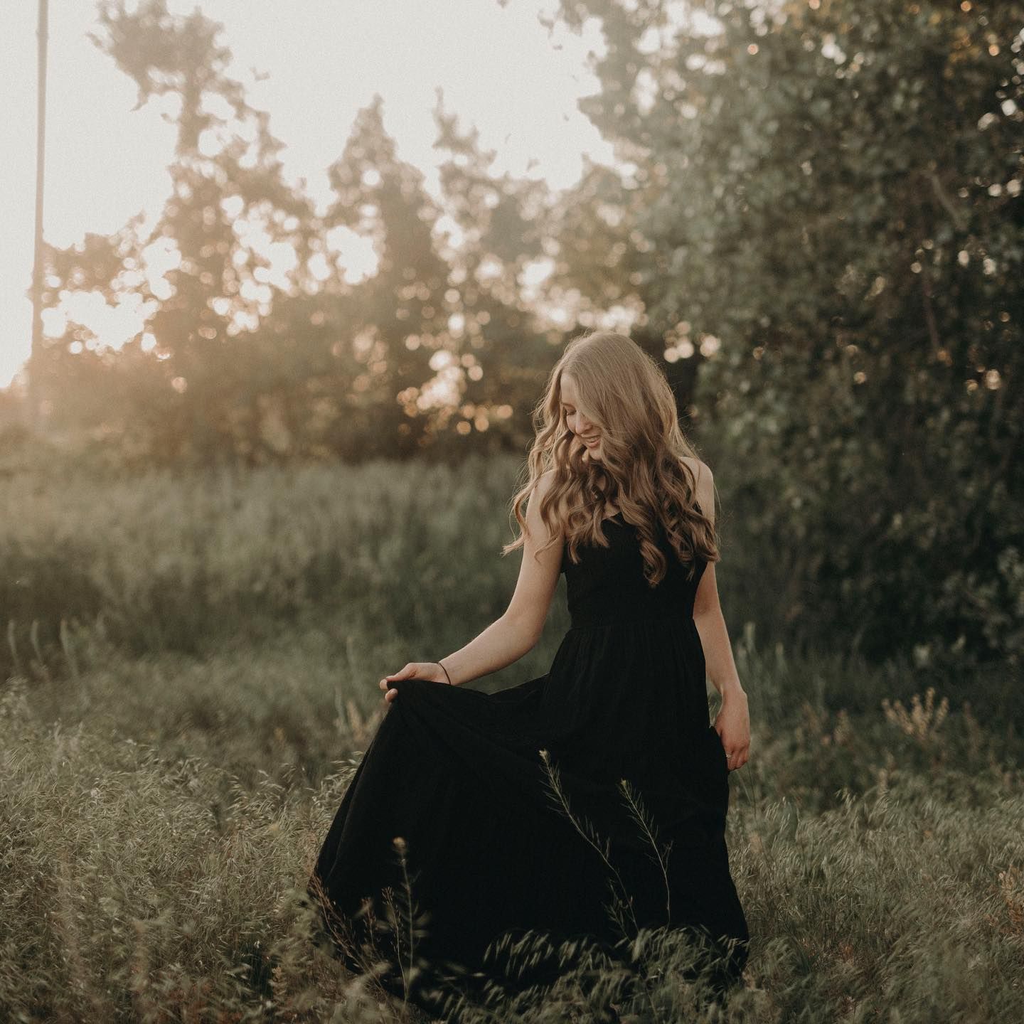 A woman in a flowing black maxi dress stands in a sunlit meadow with lush green trees in the golden hour background.
