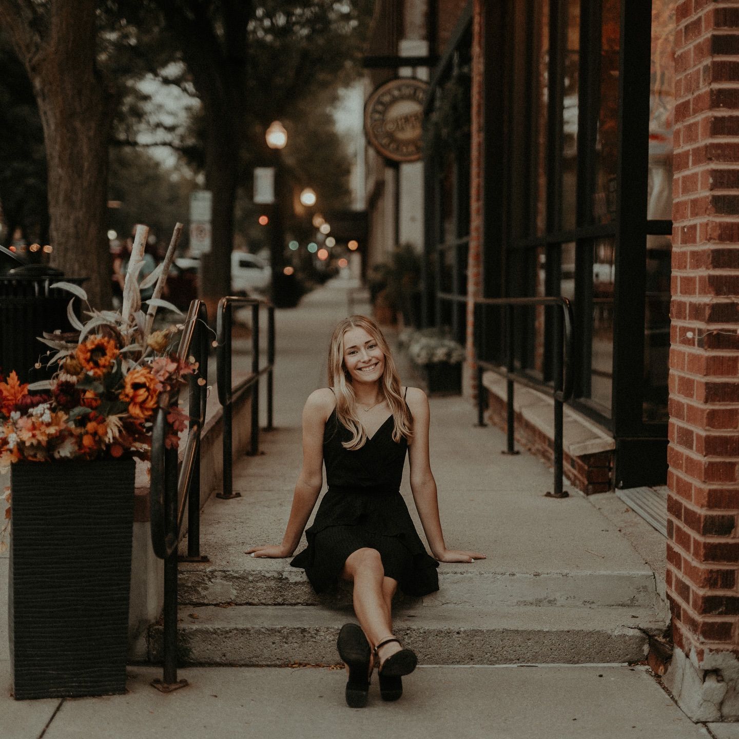 Young blonde woman in black dress sitting on steps outside a brick building with fall floral decor.