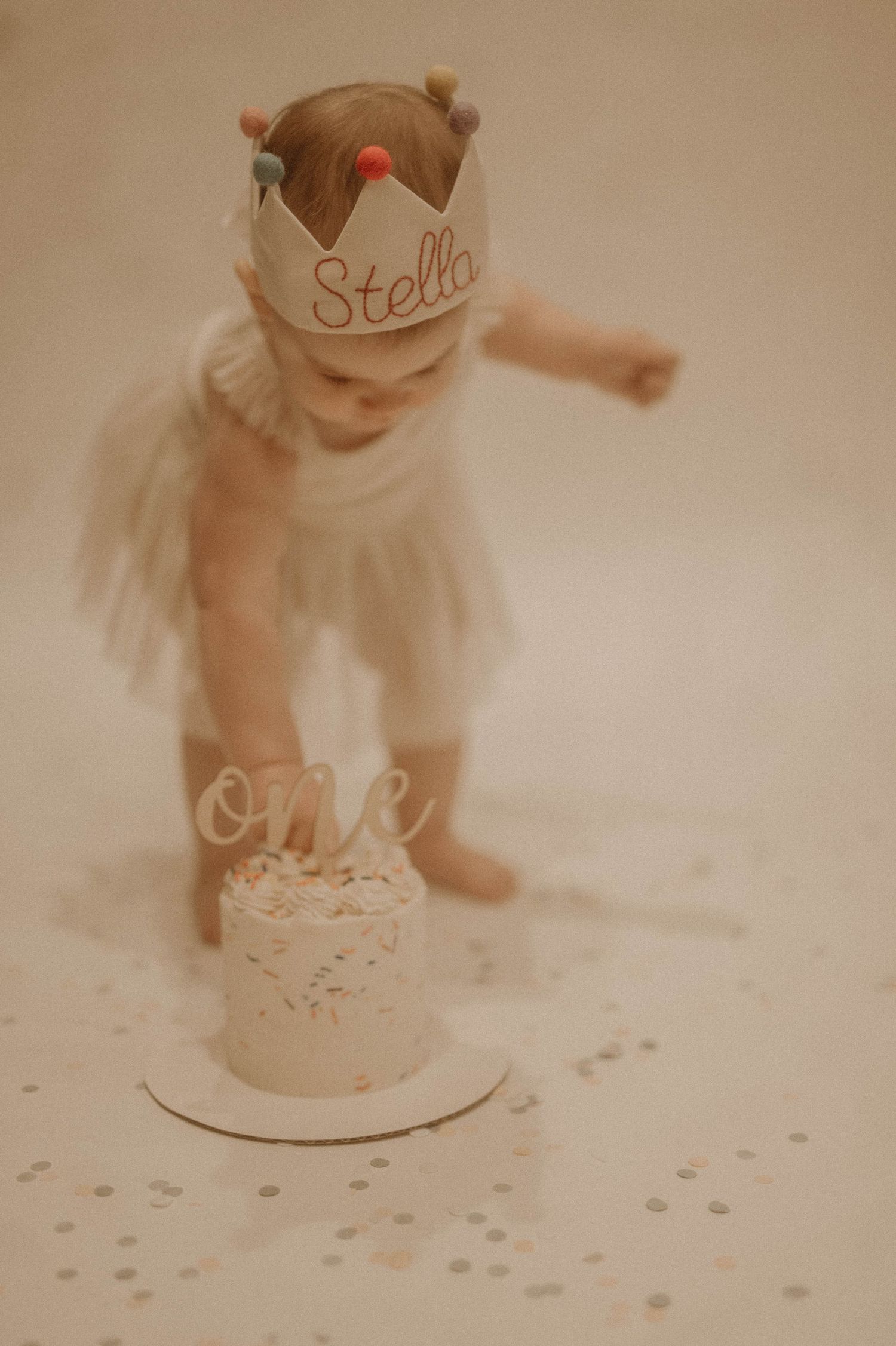 A baby in a white dress plays with confetti scattered on a light colored floor during a special celebration.
