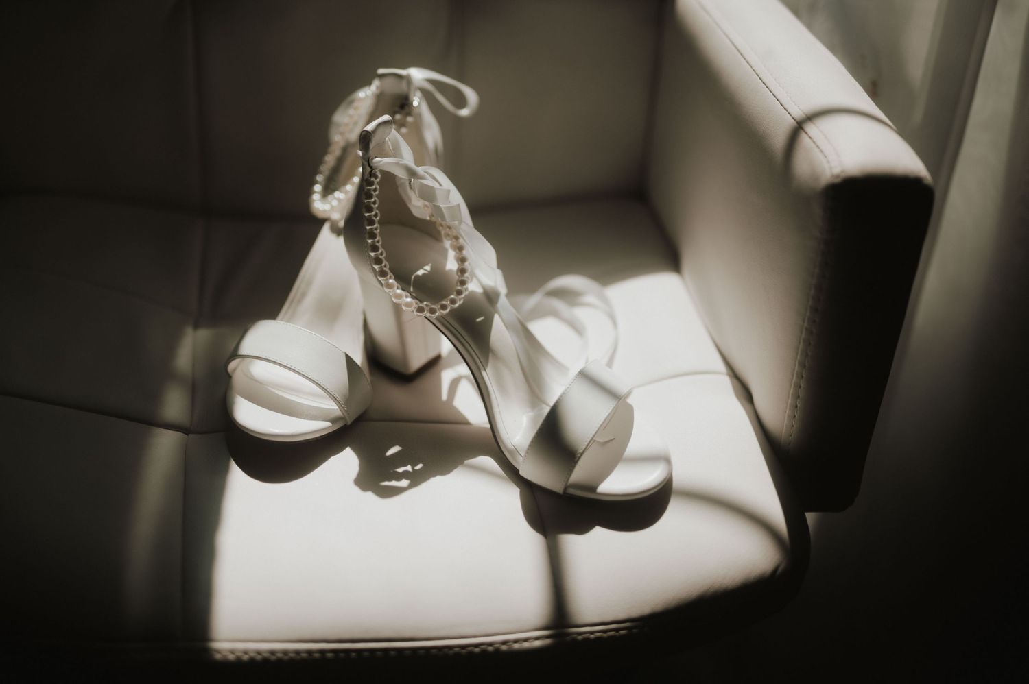 White bridal shoes and pearl necklace arranged on textured fabric with soft lighting.