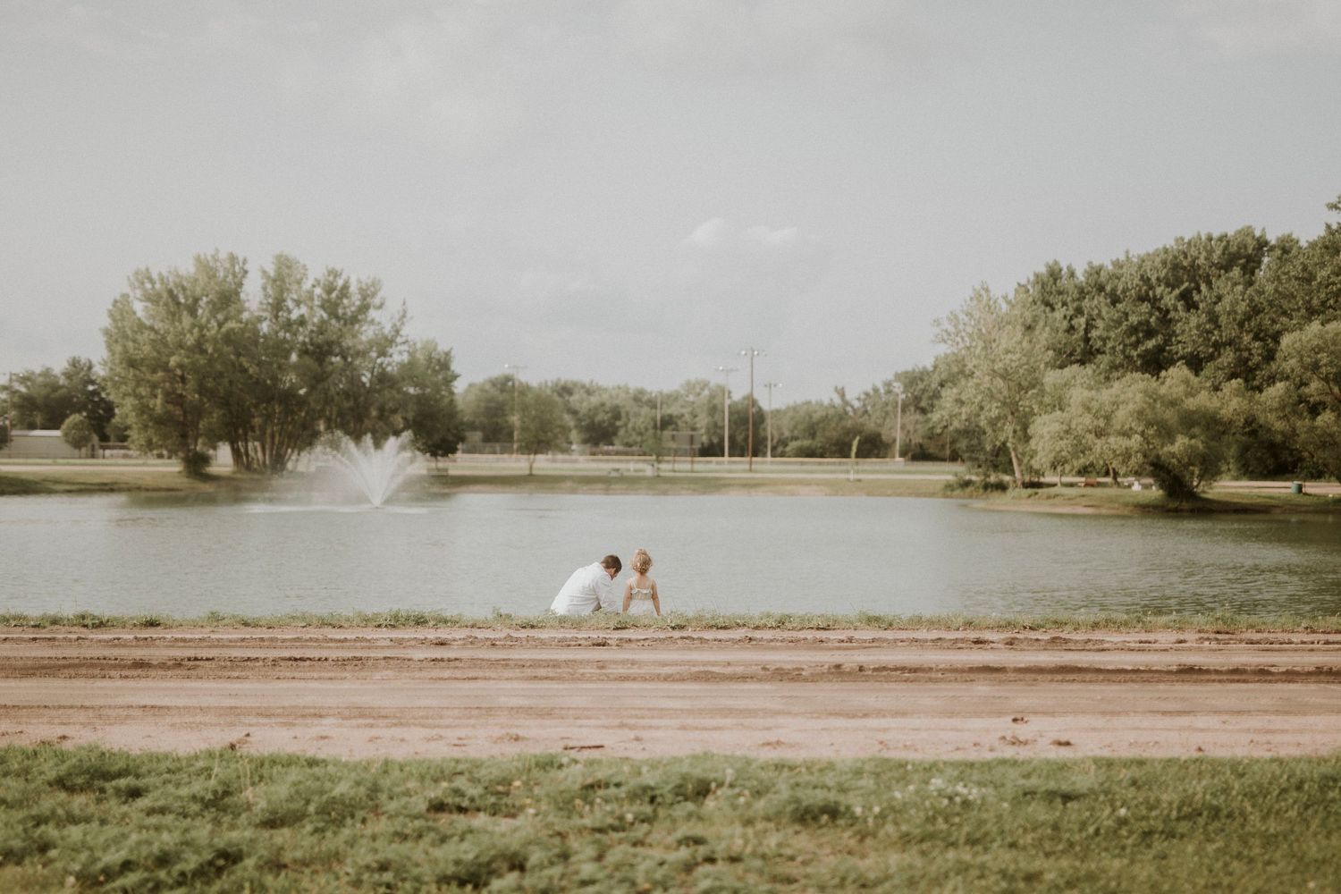 Two people sit together on a wooden dock overlooking a serene lake surrounded by trees on a sunny day.