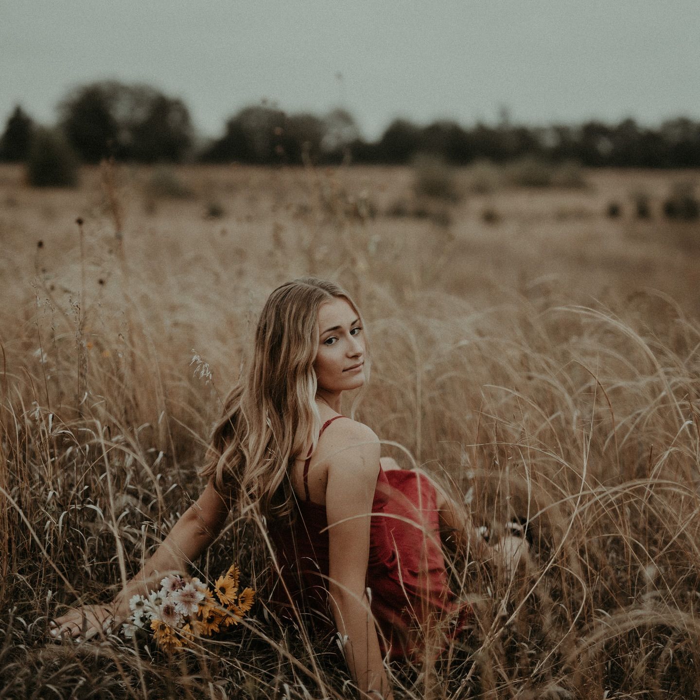 Young woman in red dress sitting in golden field holding wildflowers, with trees in background on overcast day.