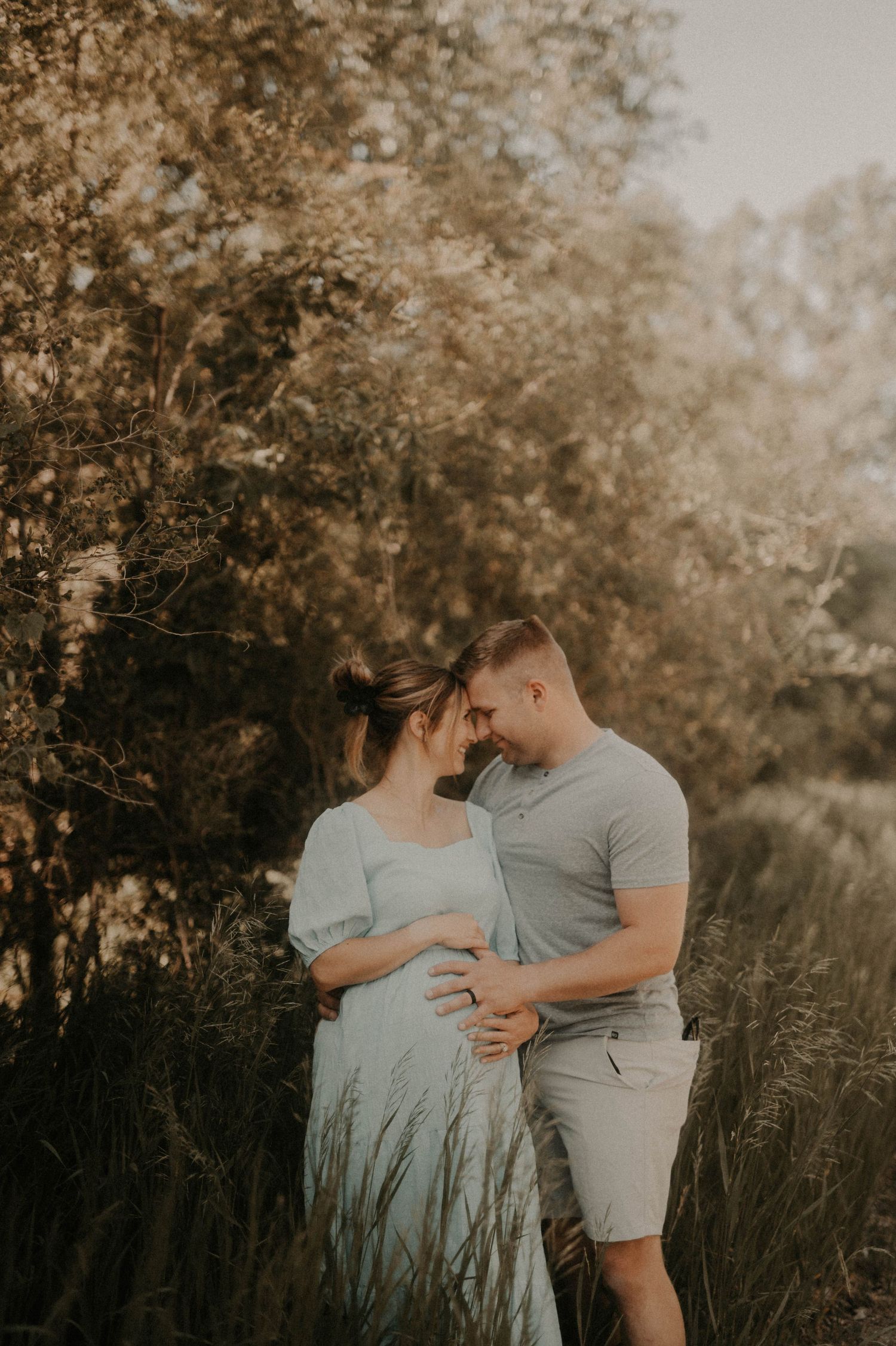Intimate maternity photo of couple embracing in tall grass during golden hour with soft sepia tones.