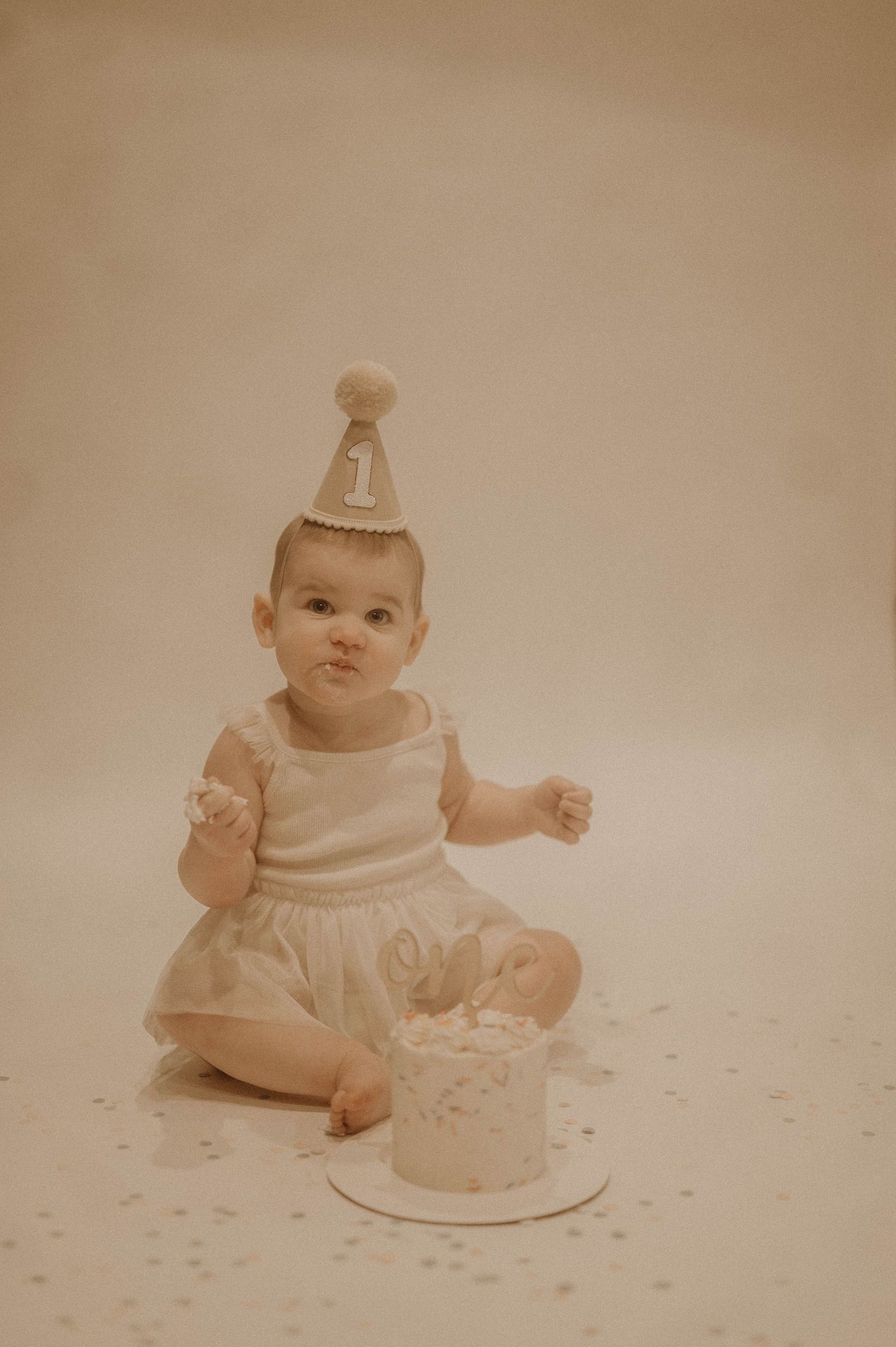 Baby in white dress sits on polka dot backdrop with birthday cake during first birthday photo session.