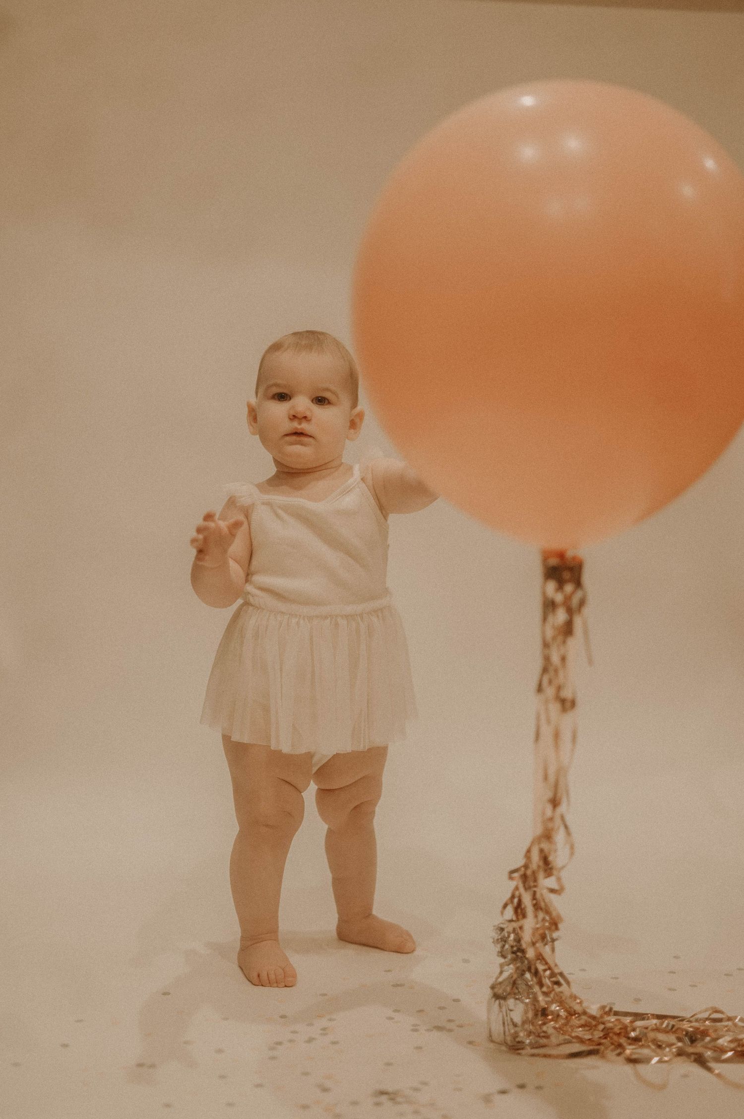Baby in white dress standing on white backdrop with peach balloons and gold streamers in vintage style photography.