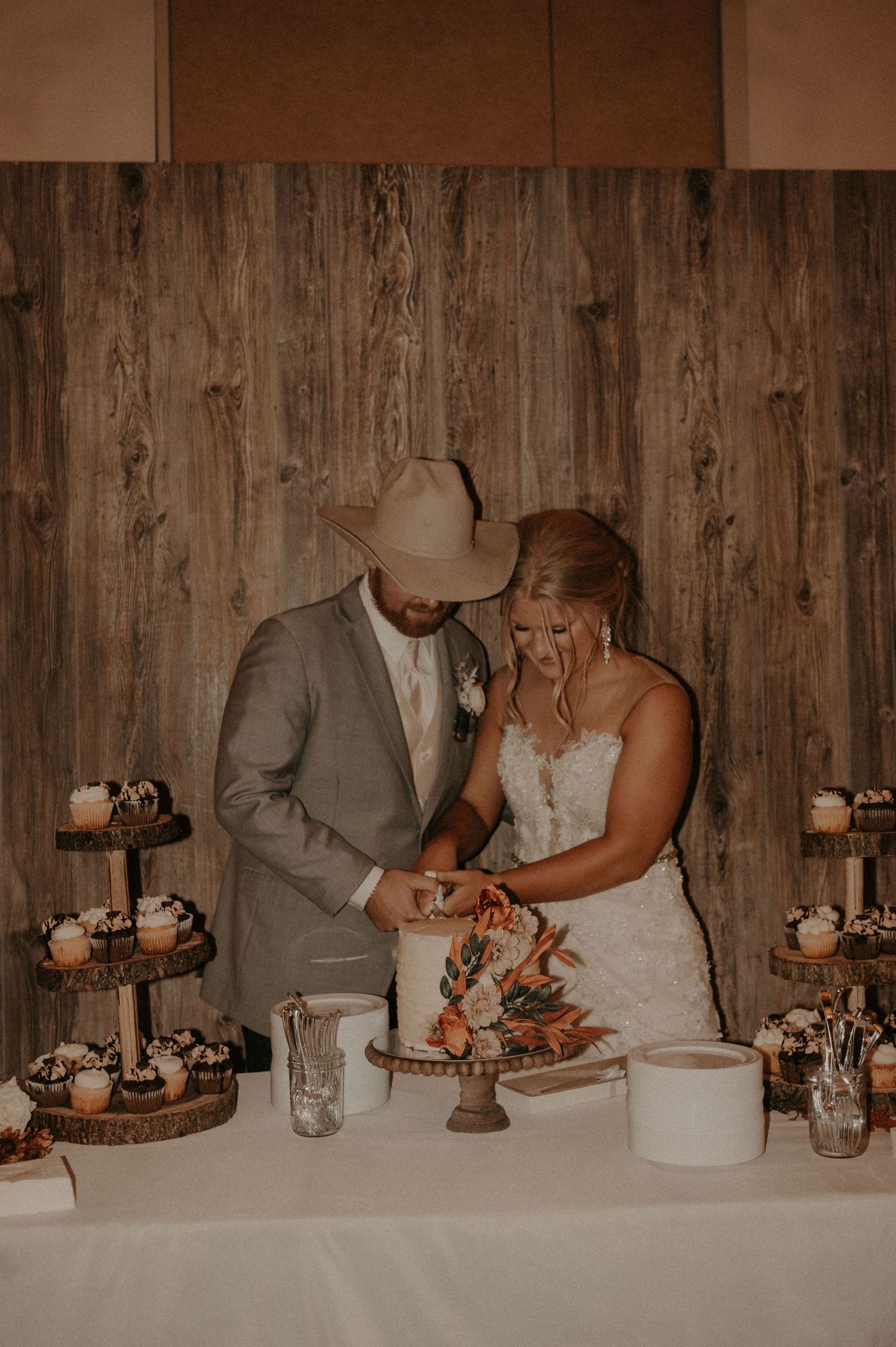 Newlyweds share an intimate moment while cutting their wedding cake against a wooden backdrop.