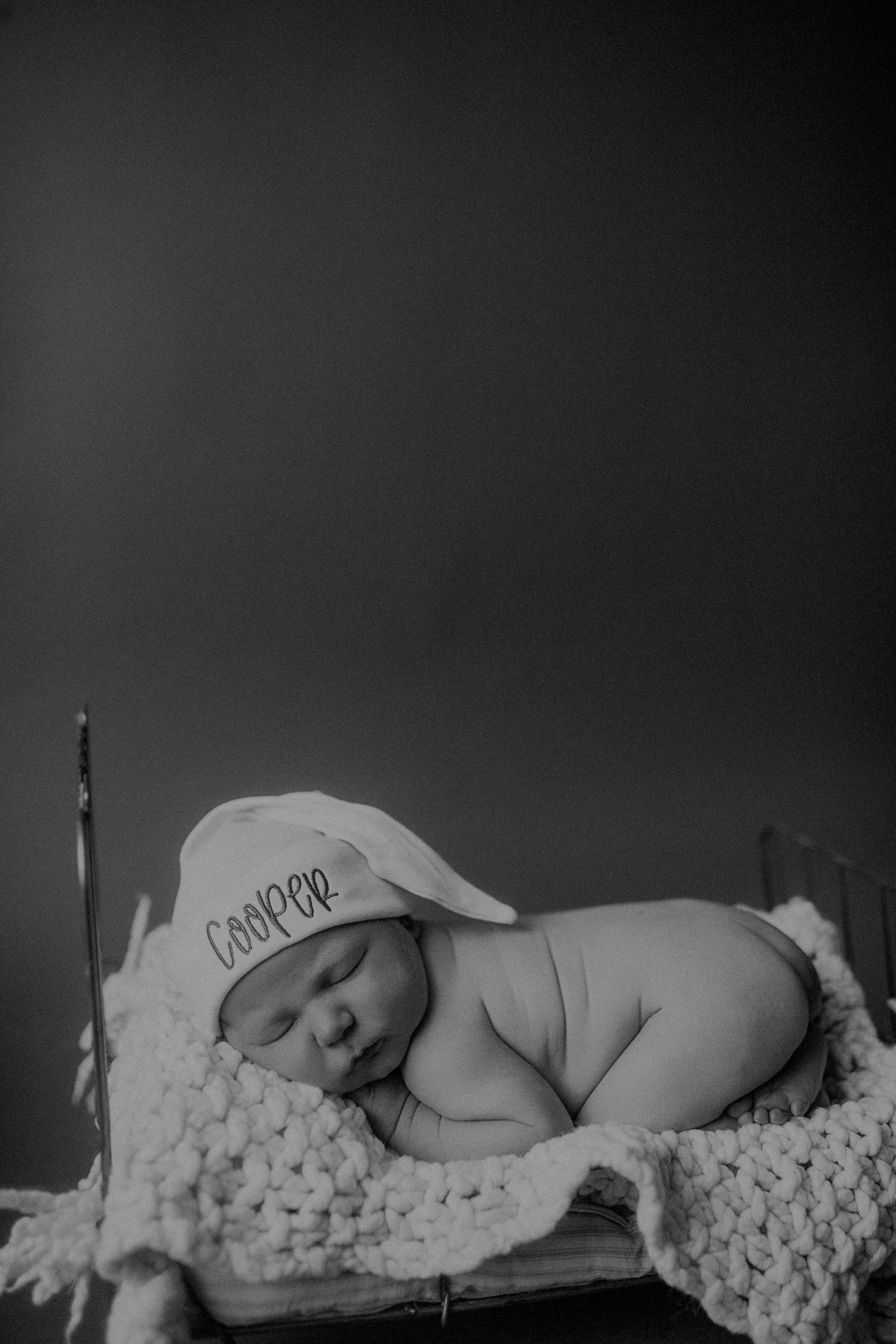 Sleeping newborn wearing a white cap on textured knit blanket in black and white.