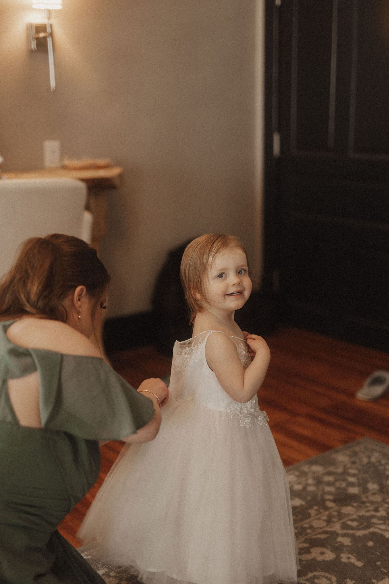 A white tulle flower girl dress twirls across a patterned carpet during wedding preparations.
