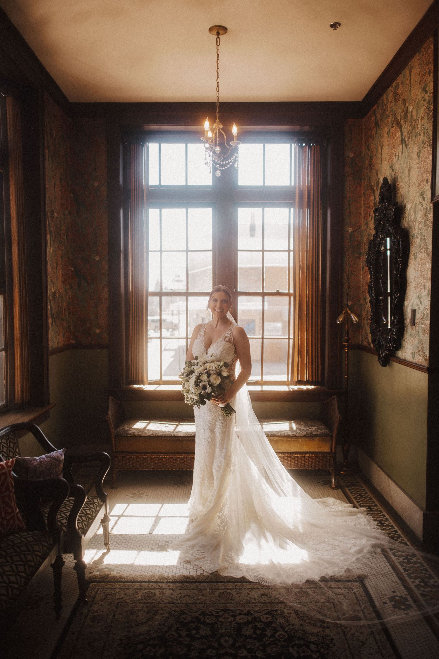 Sunlight streams through windows illuminating a bride in a flowing white gown and long veil in an elegant vintage room.