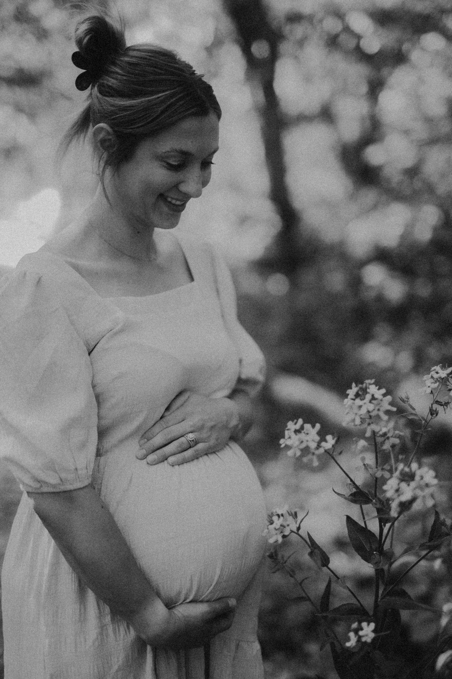 A black and white series shows a person in a light dress holding their pregnant belly near blooming flowers in a garden.