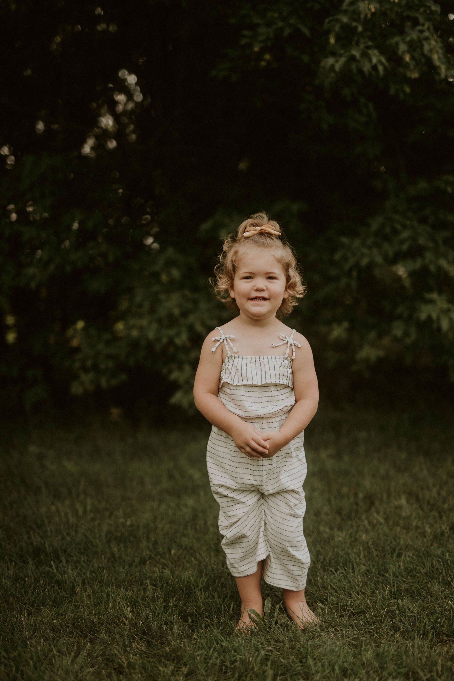 A young child stands in a grassy field wearing a light-colored romper outfit on a summer day.