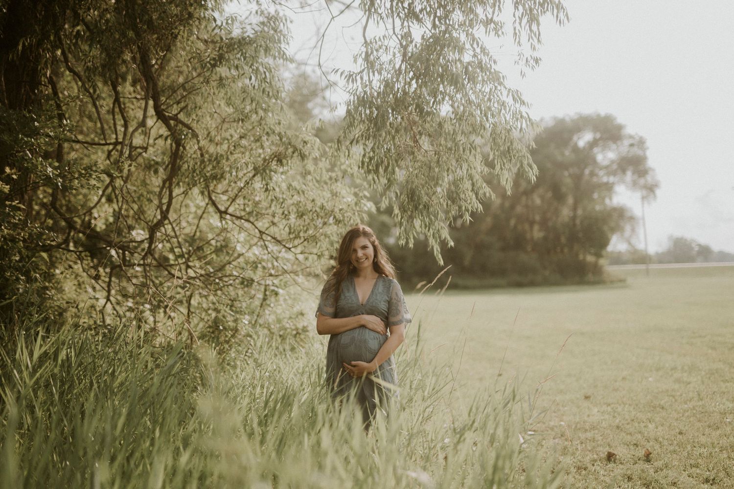 A dreamy outdoor maternity photo shoot in a sunlit field with tall willow trees and soft natural lighting.