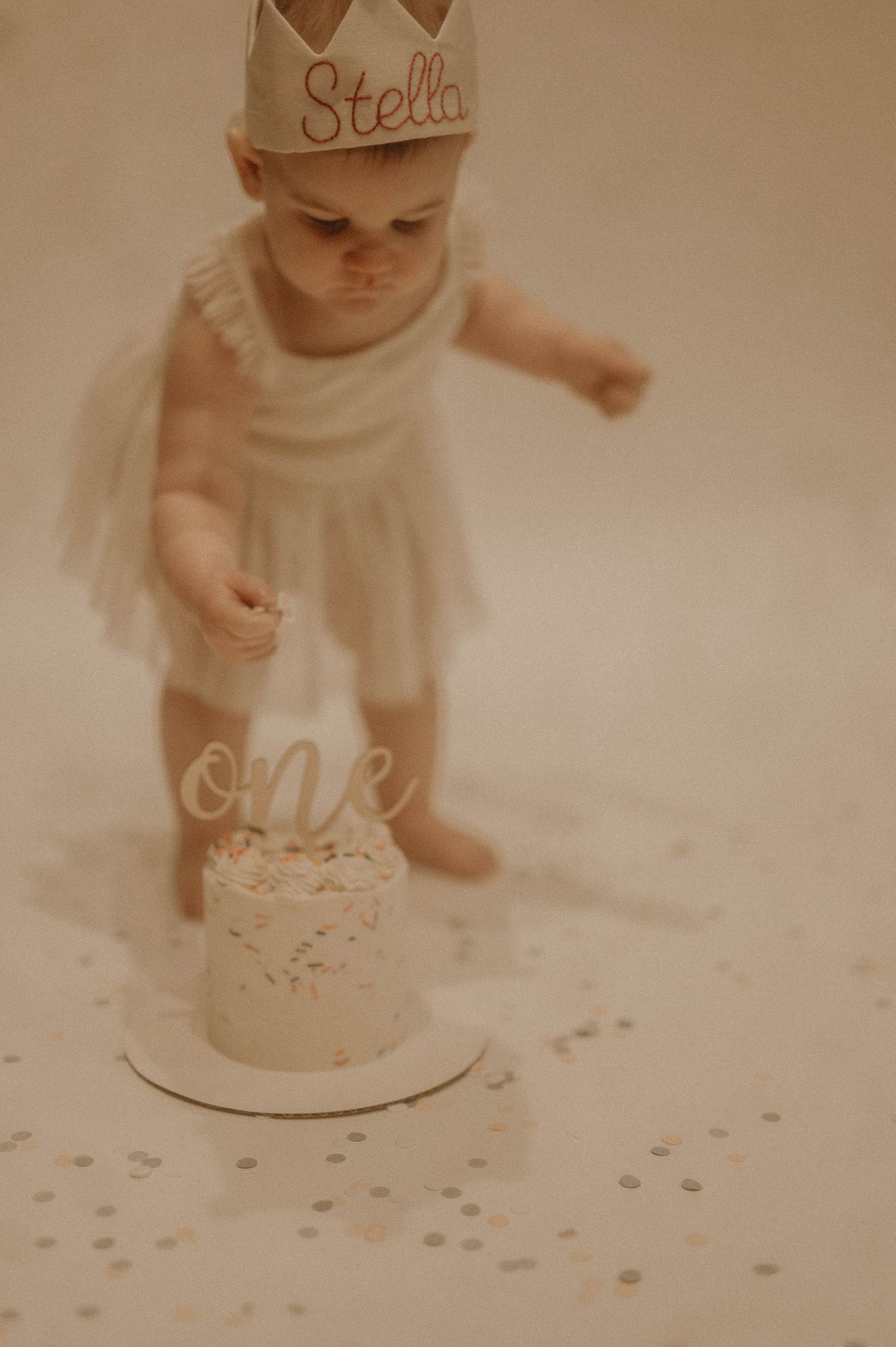 A baby in a white outfit plays with confetti and sparkles scattered on a light colored surface in a soft vintage style photo.