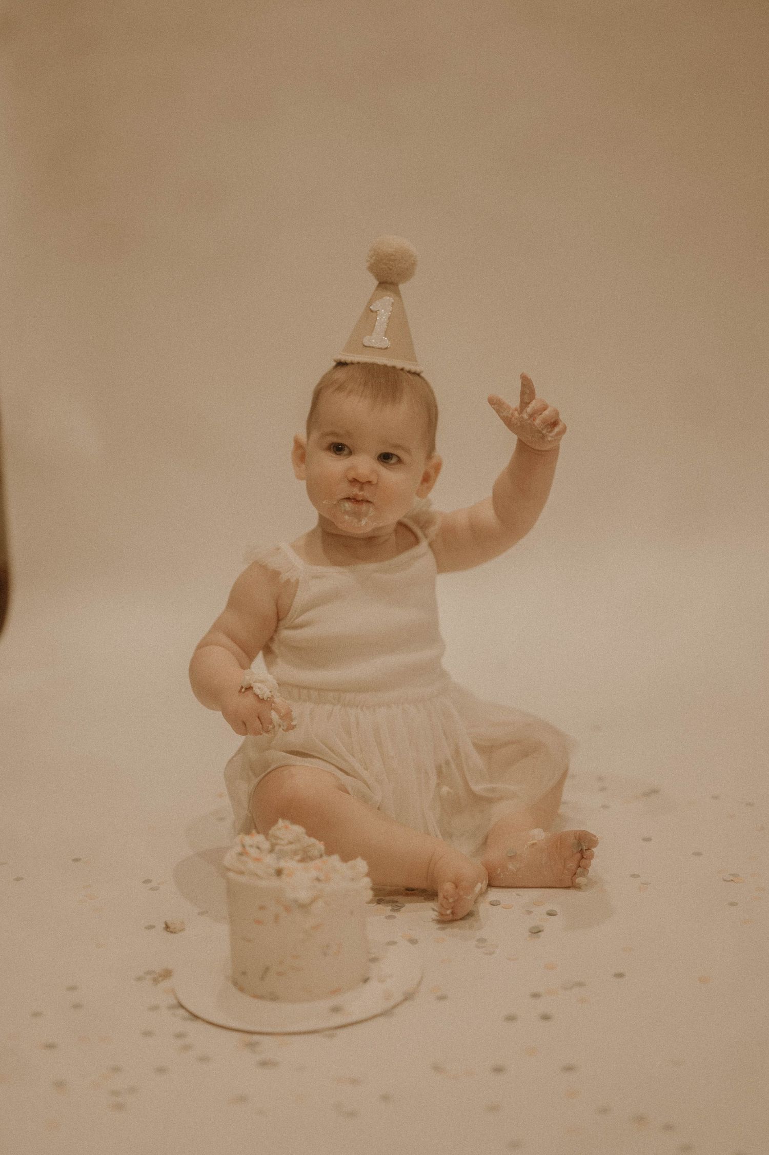 Baby in white dress sits on floor with birthday cake while confetti sparkles around in sepia-toned photo series.
