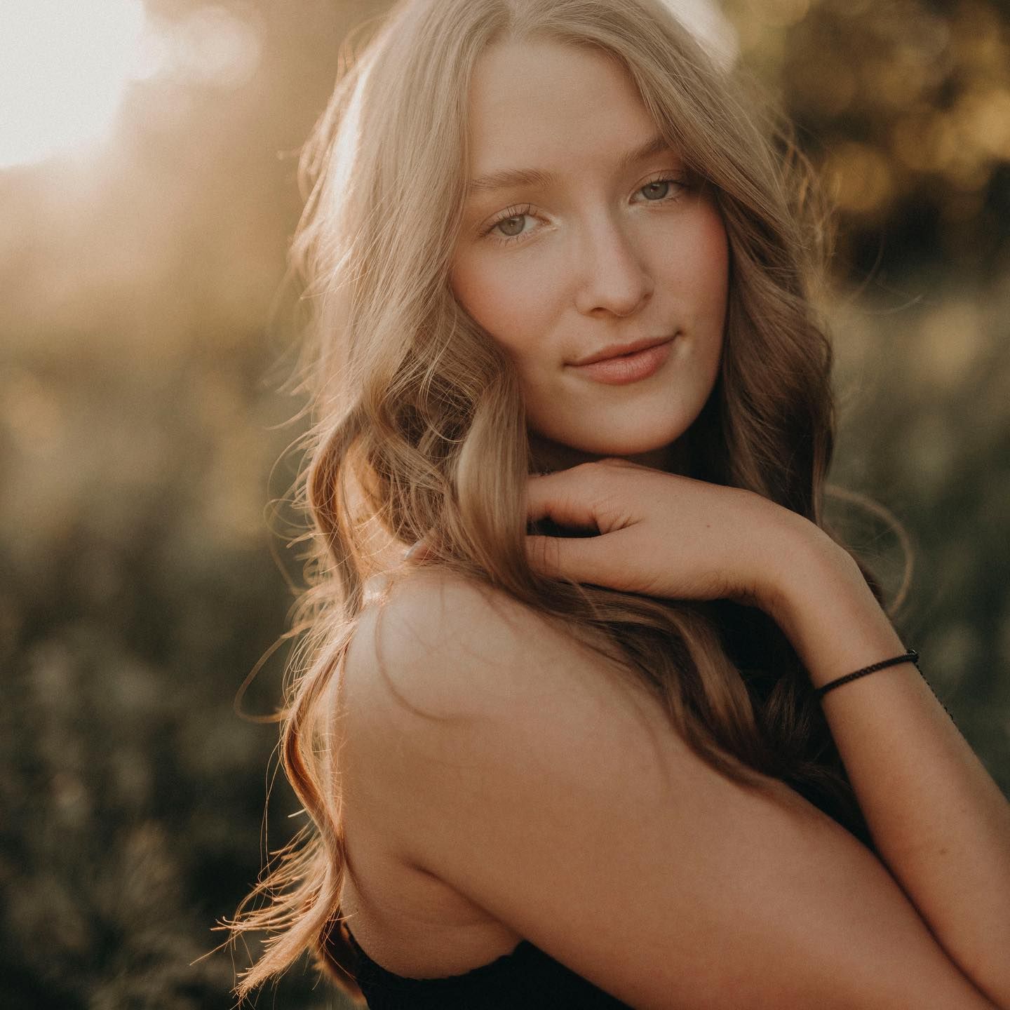 Young woman with long blonde hair poses outdoors in golden sunset light, wearing a black top with a bracelet.