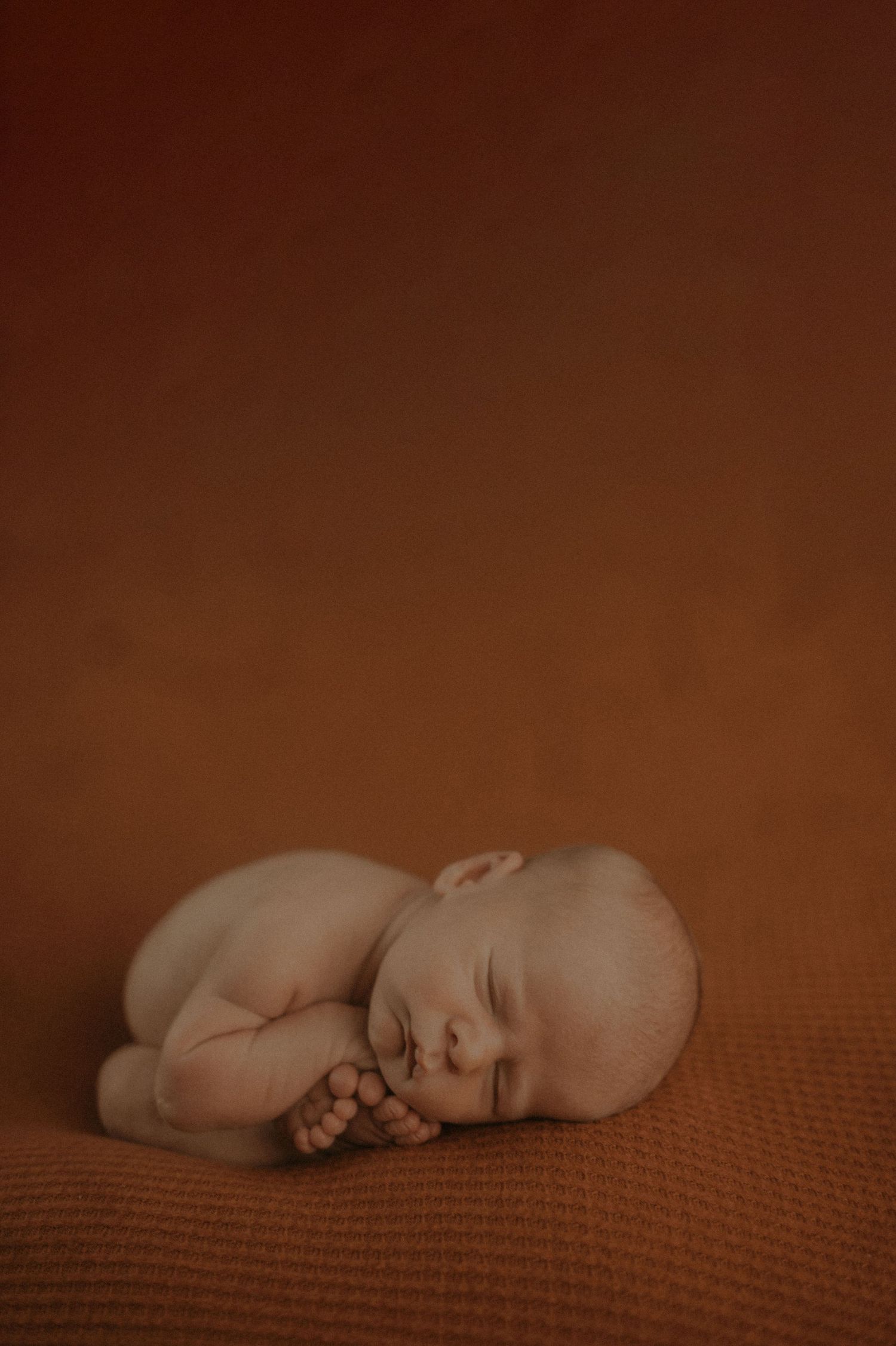 Sleeping newborn baby in natural pose on brown textured blanket with warm lighting.