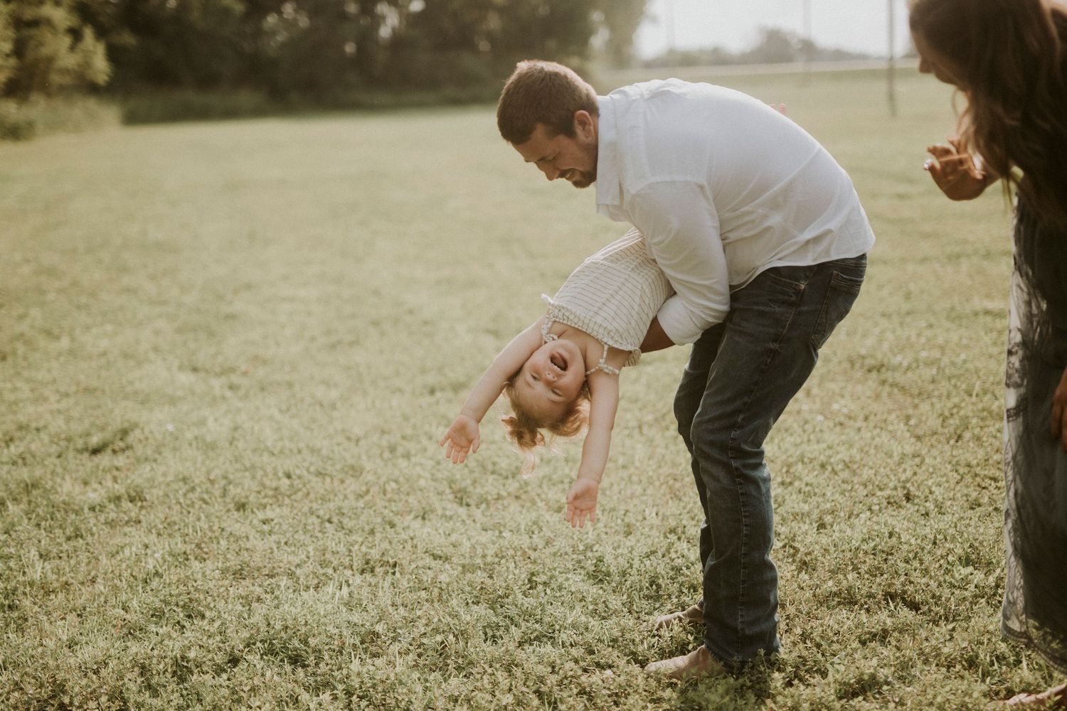 A toddler in a white sweater and gray pants plays in a grassy field on a sunny autumn day.
