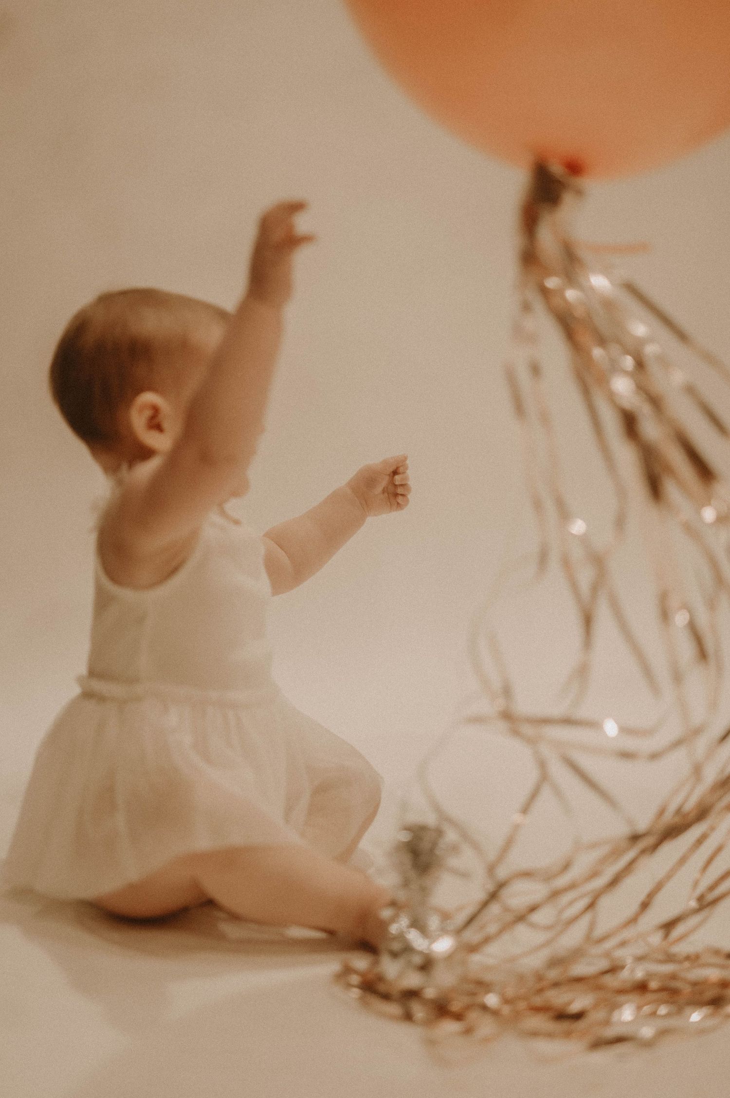 A sepia-toned photo series showing a baby in a white dress reaching upward while sitting against a soft background.