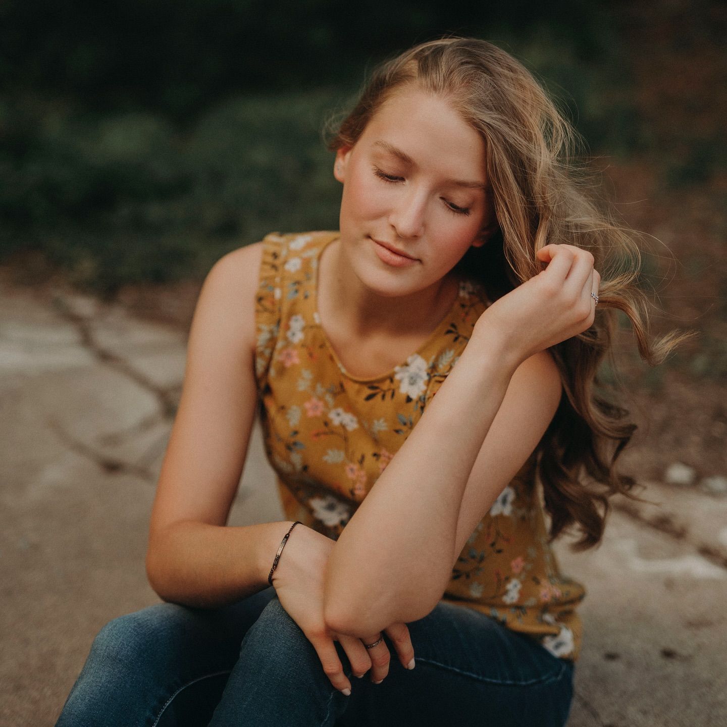 Young woman in floral top and jeans sits outdoors, gazing down thoughtfully with wavy hair flowing over shoulder.