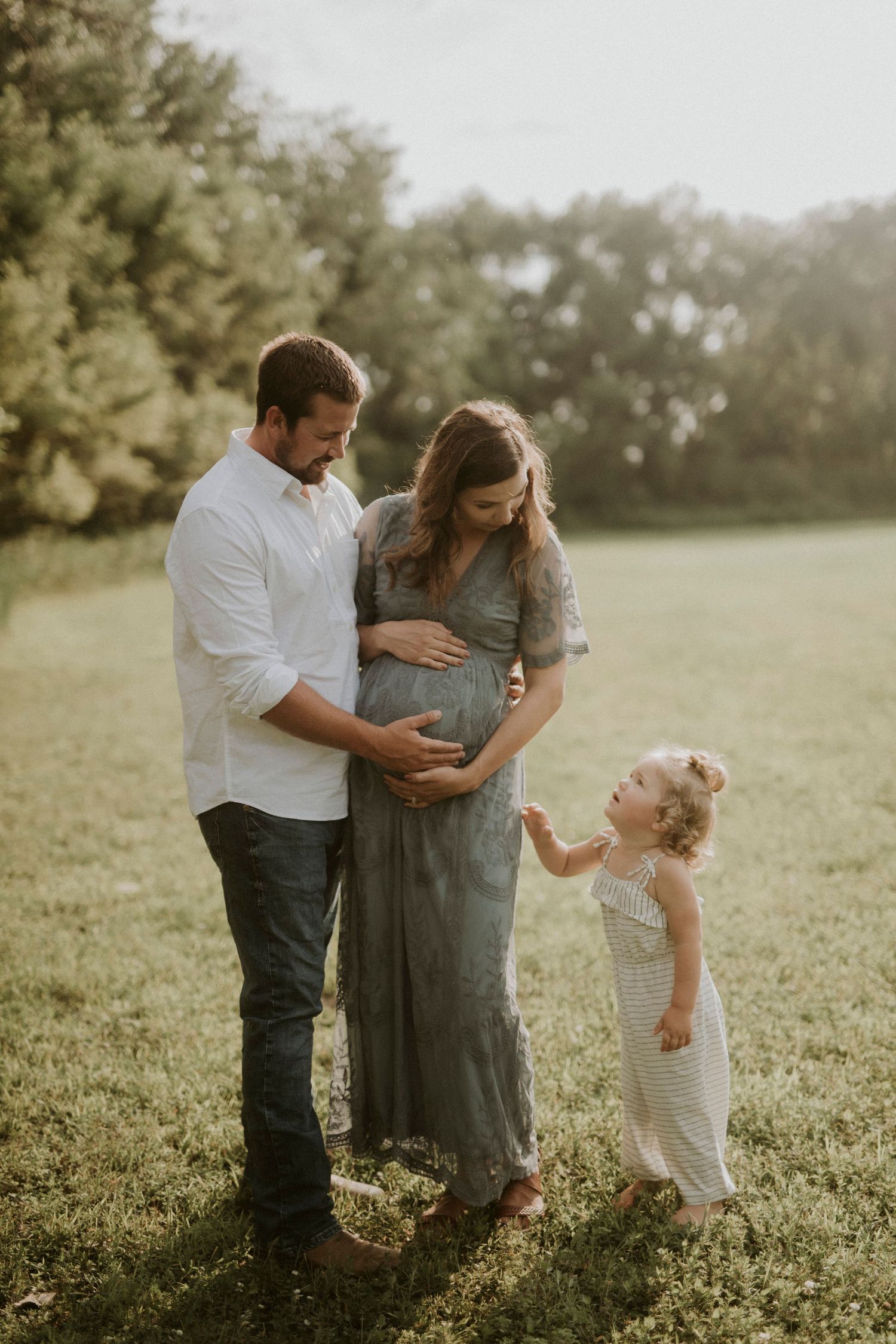 A family shares a tender moment in a sunlit field as they await their new arrival, with a toddler in a white dress nearby.