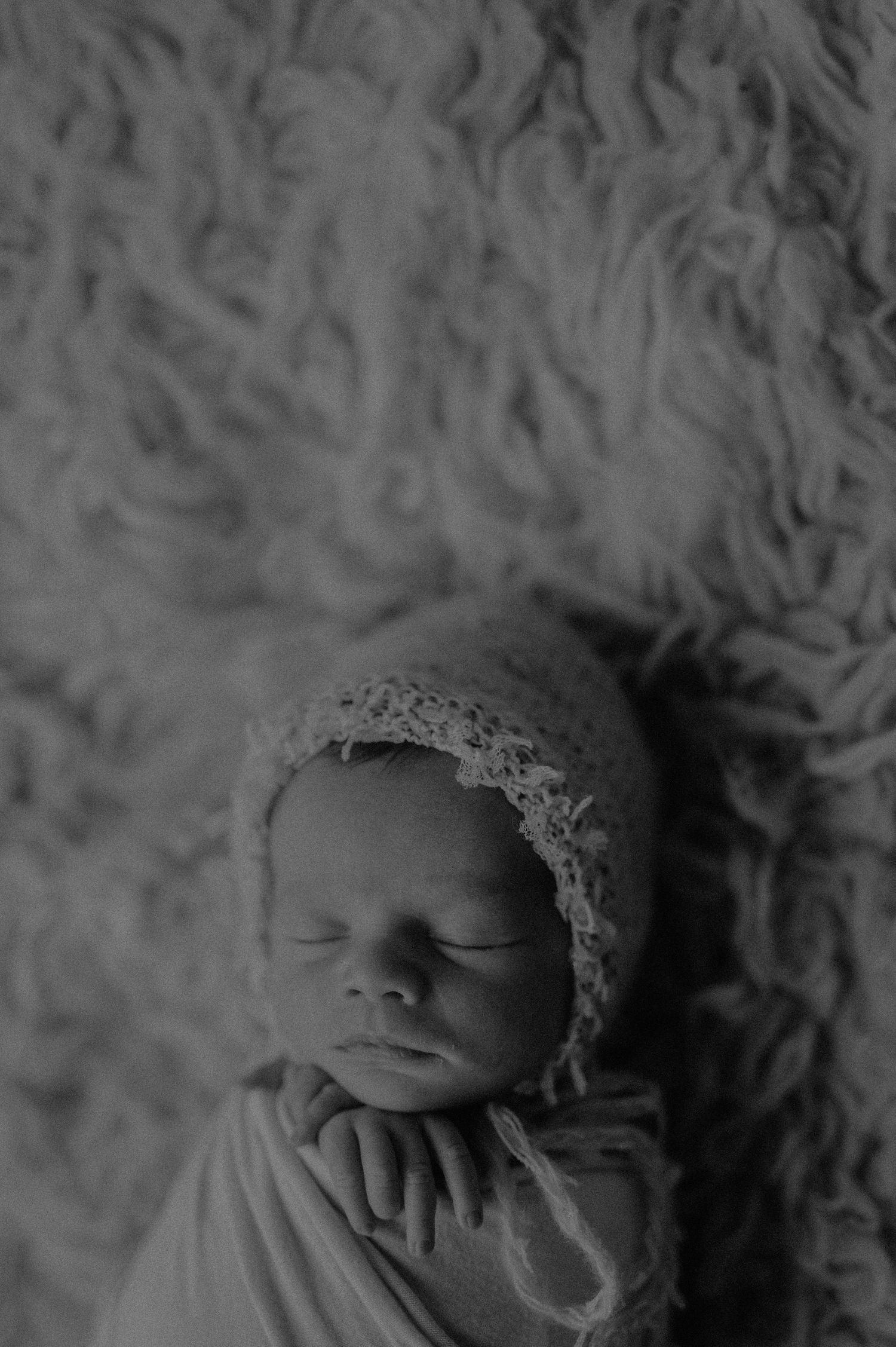 Monochrome portrait of sleeping infant wearing lace headpiece on textured backdrop.