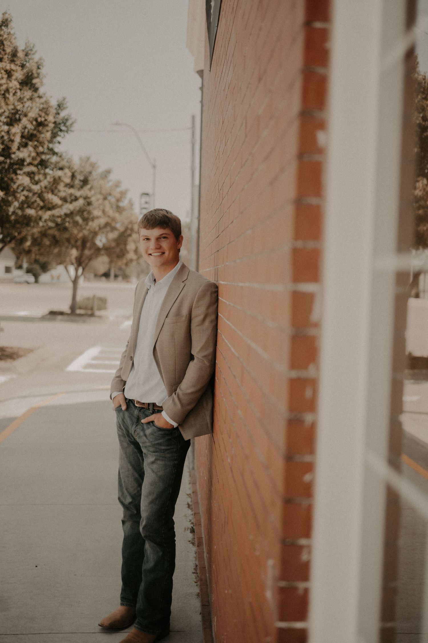 A person in a gray suit and dark pants stands against a brick wall in warm natural lighting.