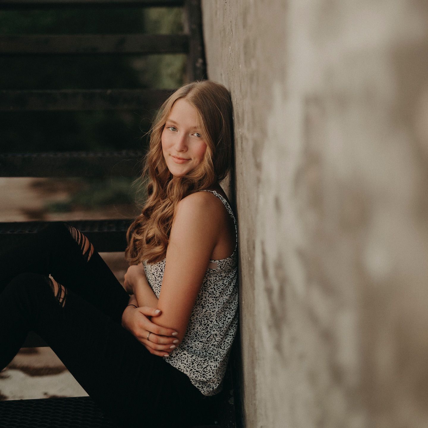 Young teenage girl with wavy blonde hair sitting against a concrete wall on steps, wearing a floral tank top.