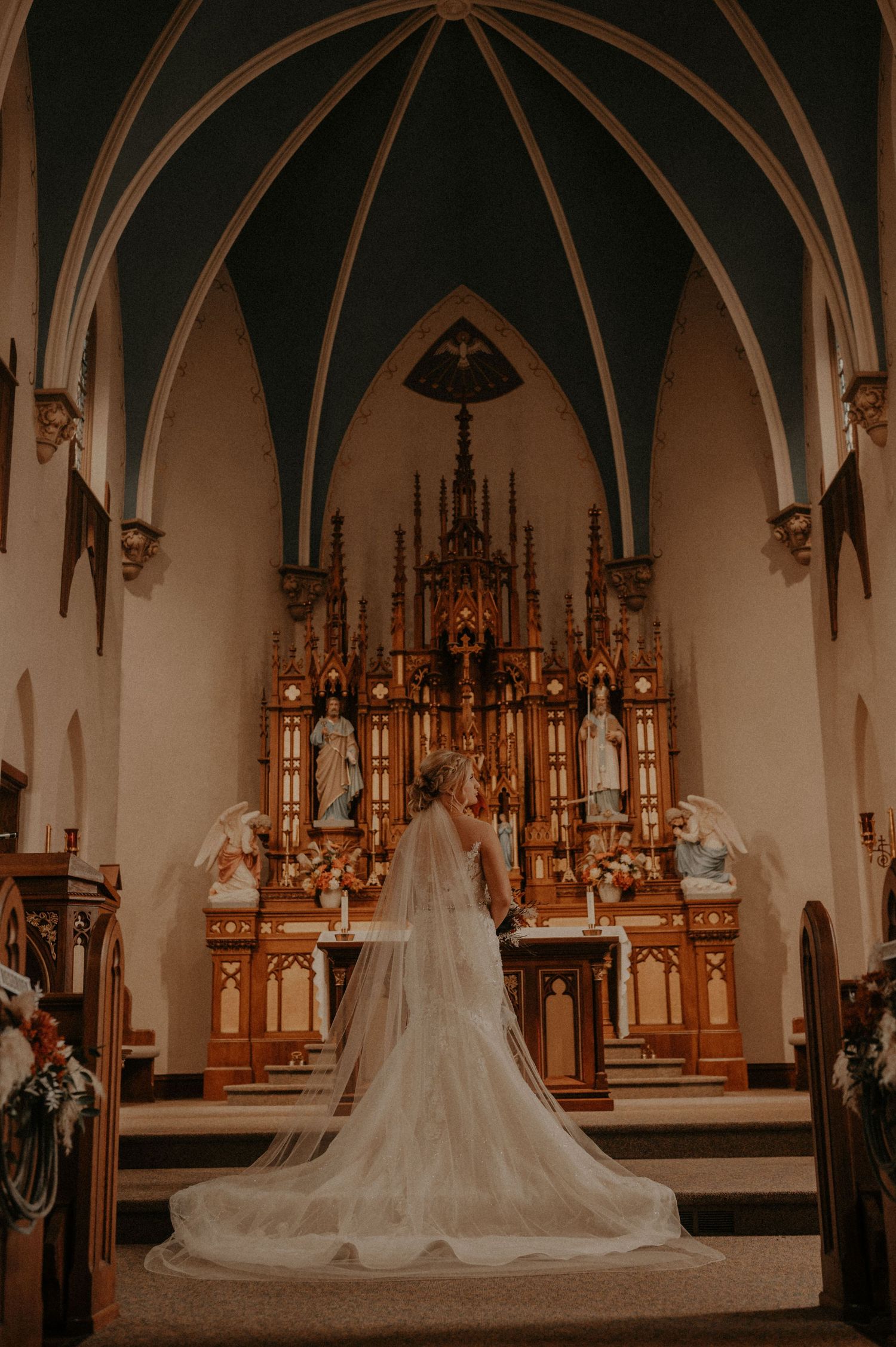 Bride in flowing white gown and veil stands before an ornate golden church altar.