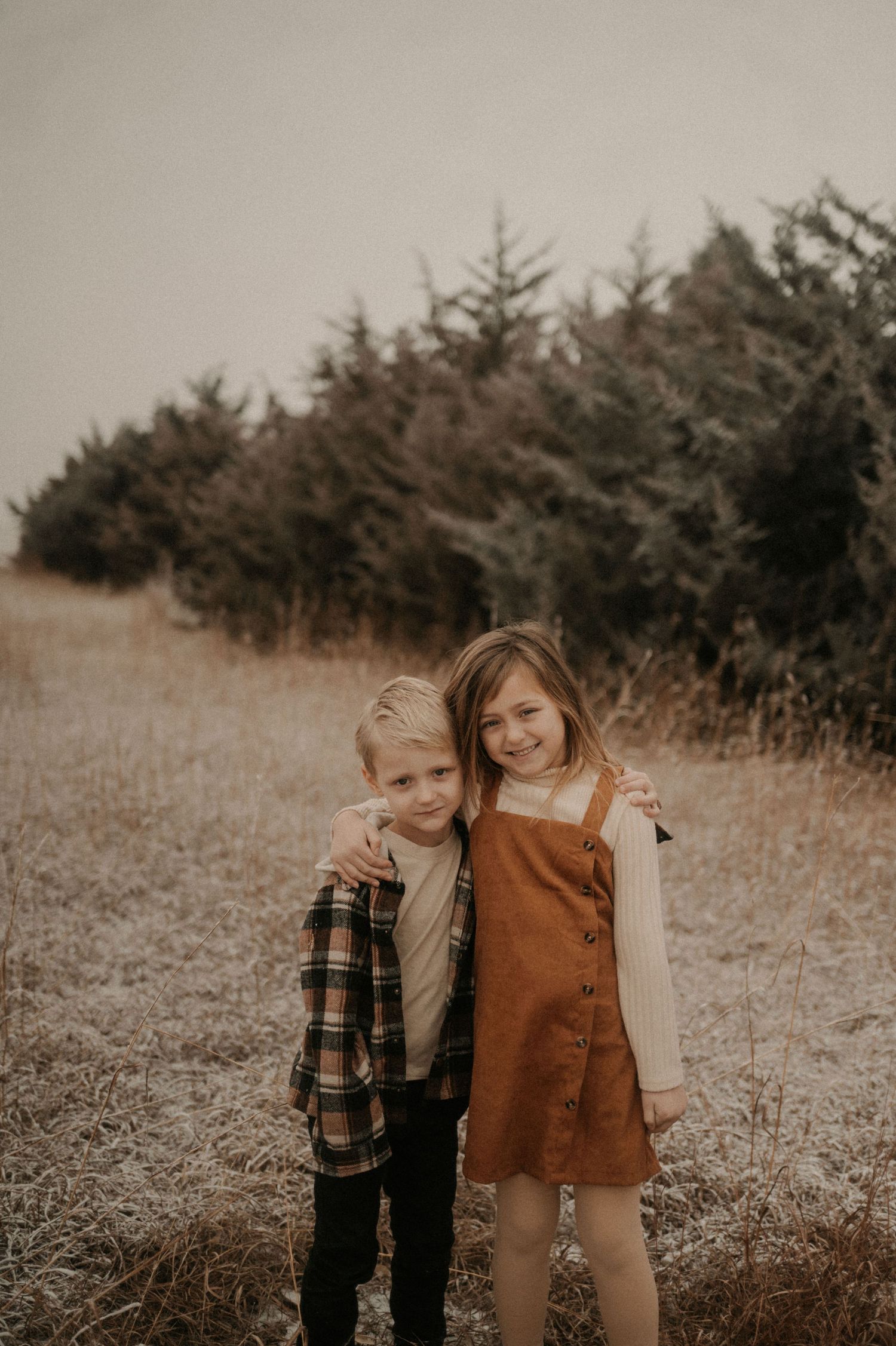 Two children stand together in a field with evergreen trees in the background during golden hour.